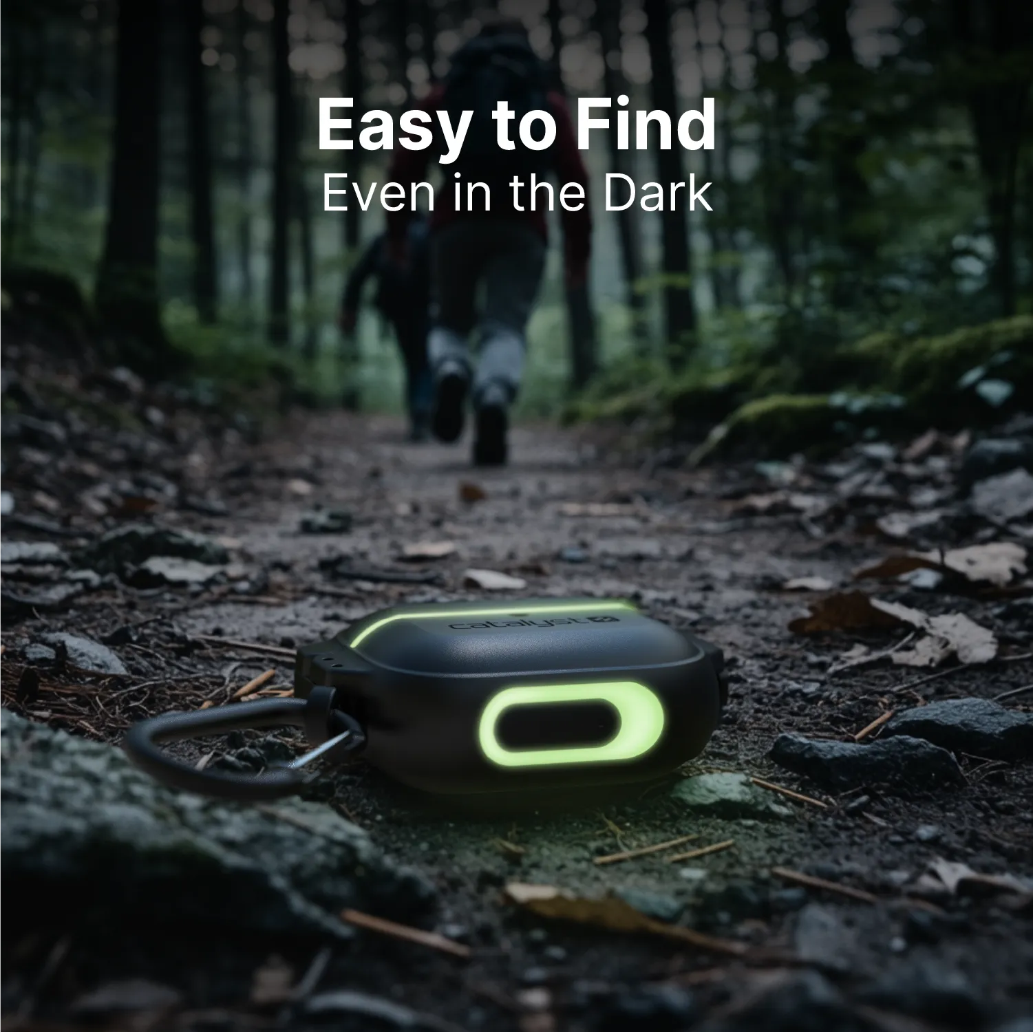 A rugged black Catalyst Case US AirPods Pro (Gen 2/1) total protection case with green glowing edges and a carabiner rests on a forest path; someone walks away in the dim light. Text above: “Easy to Find Even in the Dark.”.