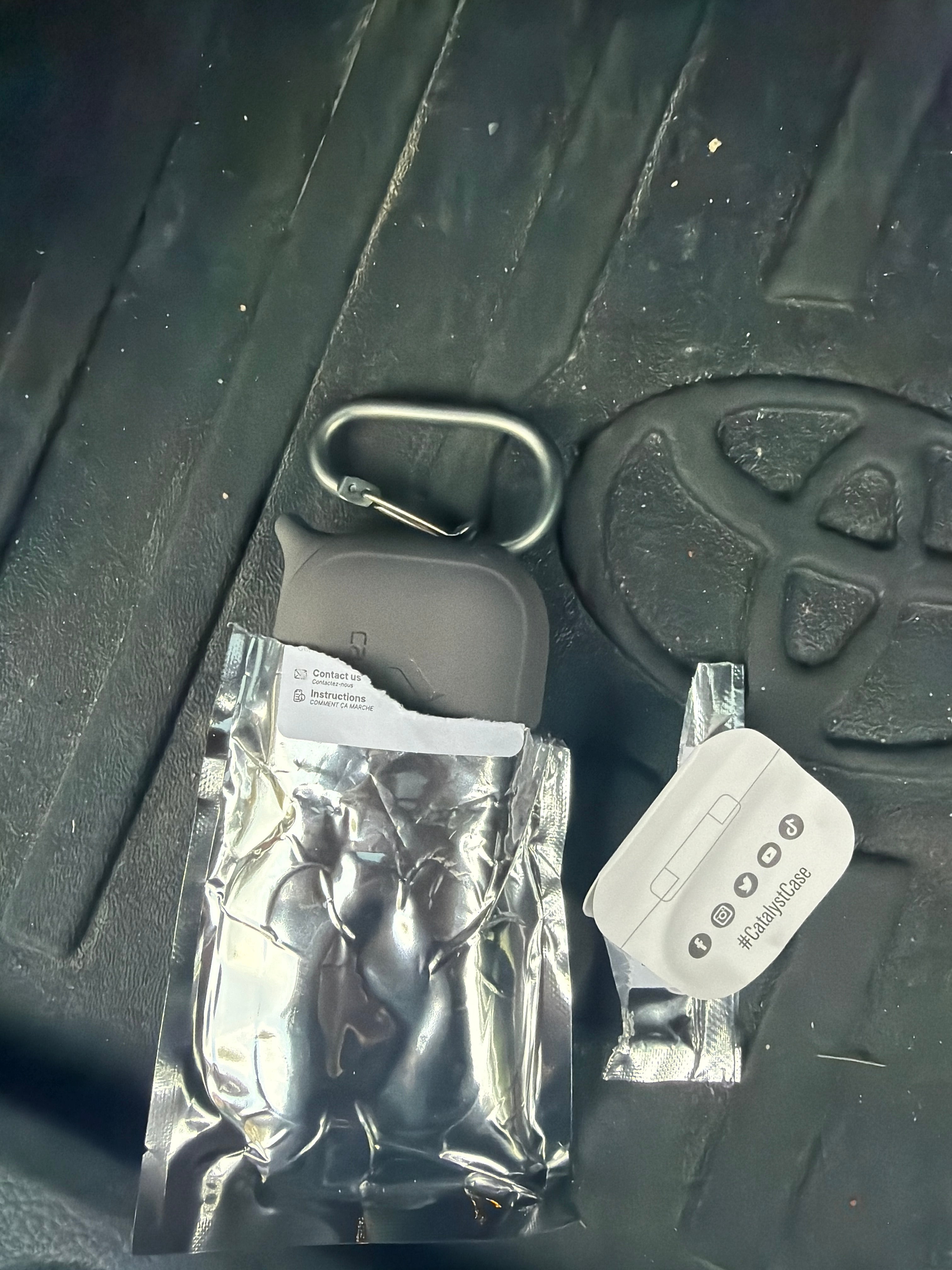 A Catalyst Case US AirPods (Gen 3) - Waterproof Case + Carabiner in Space Gray sits on a crinkled foil wrapper alongside a small white instruction sheet. All items are placed on a black car floor mat featuring a raised Toyota logo.