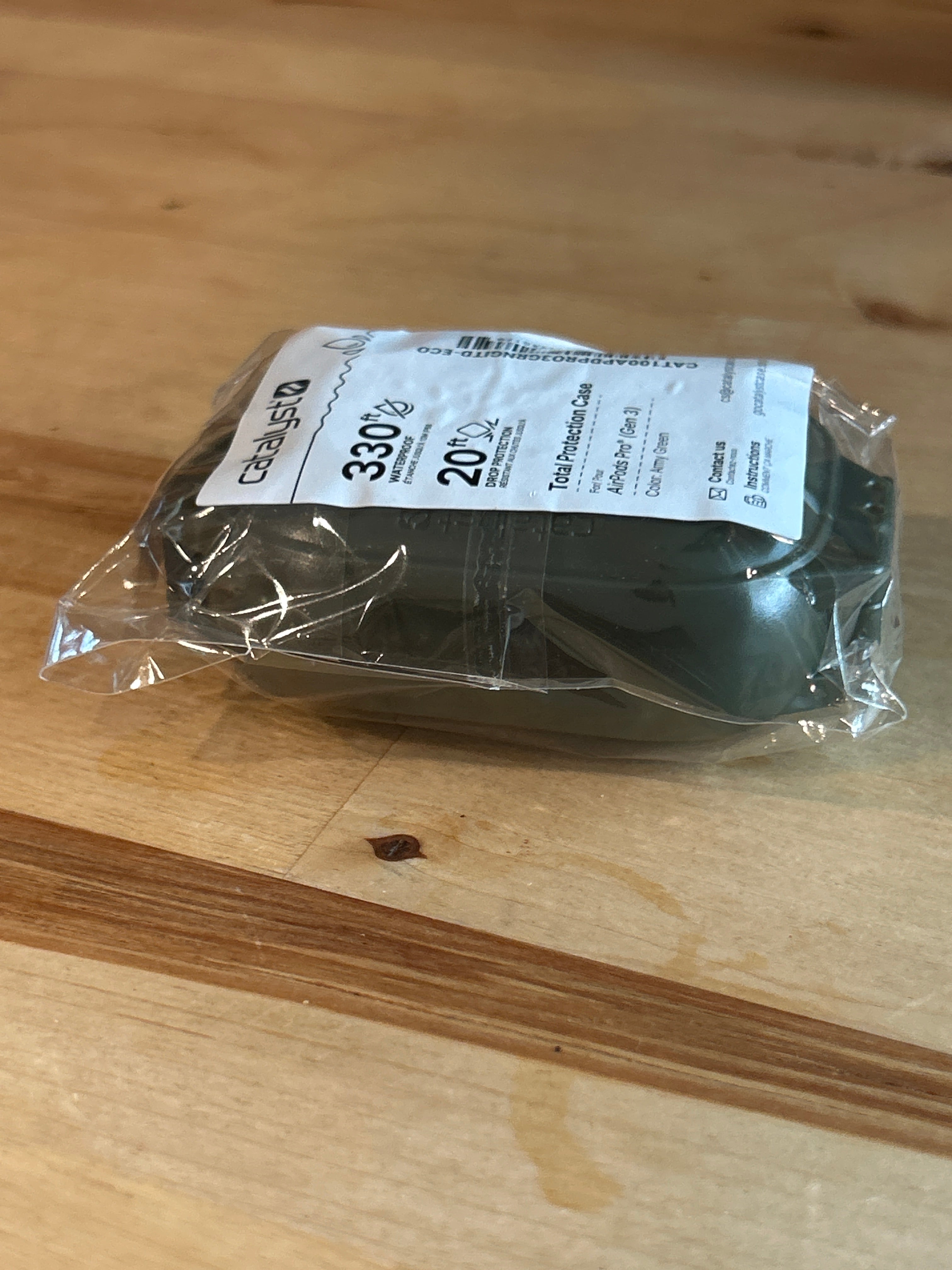 A compact, green glow-in-the-dark AirPods Pro 3 case from Catalyst Case US rests on a wooden table, its sleek, minimalist design reminiscent of a modern soap bar. The case is wrapped in clear plastic and topped with a white label featuring details like “330 g” and “20%.” Light wood with visible grain and knots forms the background.