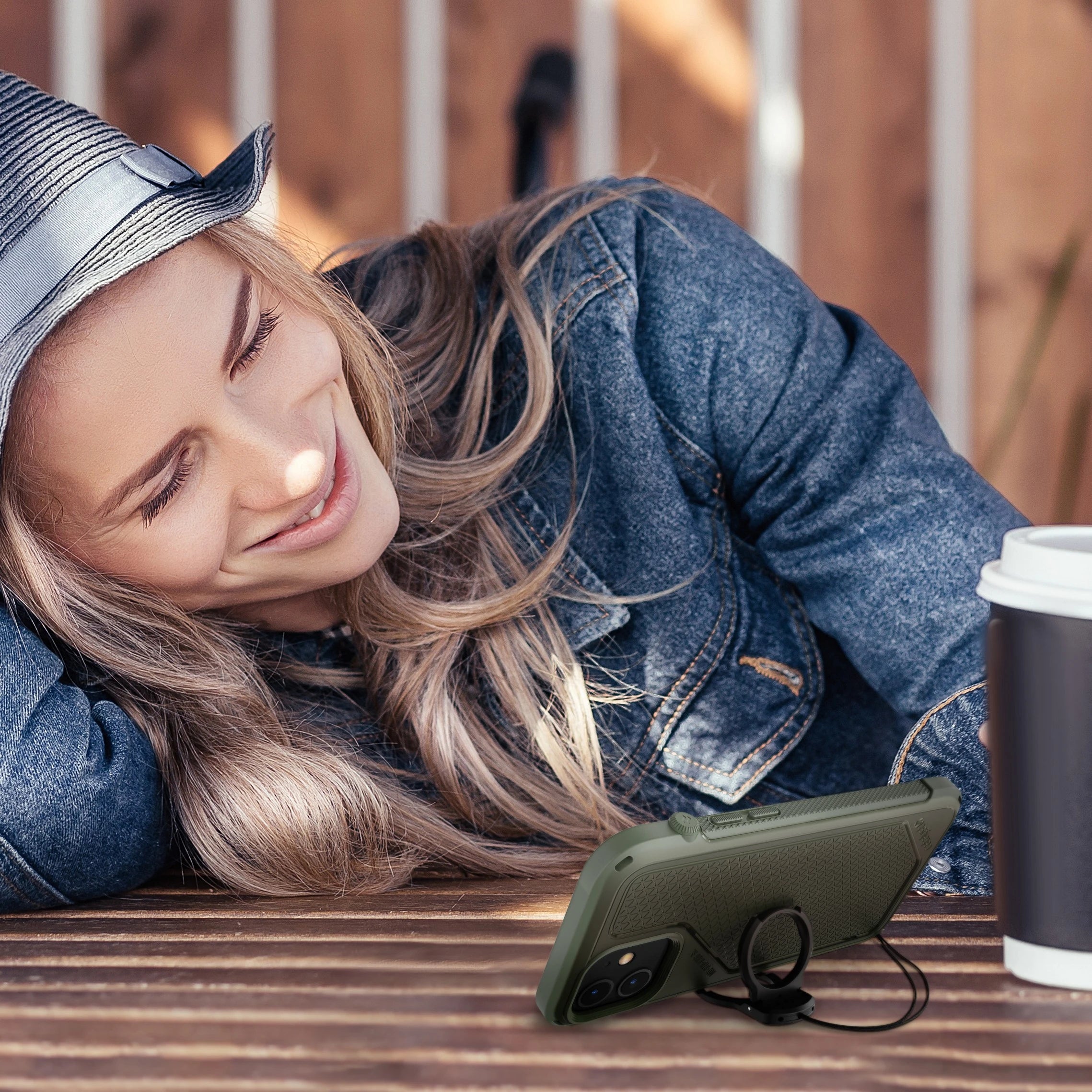 A smiling woman in a gray hat and denim jacket rests her head on a wooden table. In front of her is a Catalyst Case US iPhone 13 Pro Max - Vibe Case (MagSafe compatible), propped with a ring stand, and a white takeaway coffee cup.