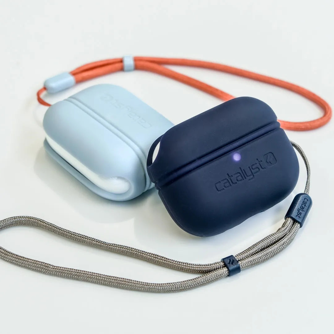 Two Catalyst Case US AirPods Pro (Gen 2) - Essential Case + Lanyard rest on a white surface: one is light blue with an orange lanyard, the other dark blue with gray. Both display Catalyst logos and have soft, rounded designs for protection.