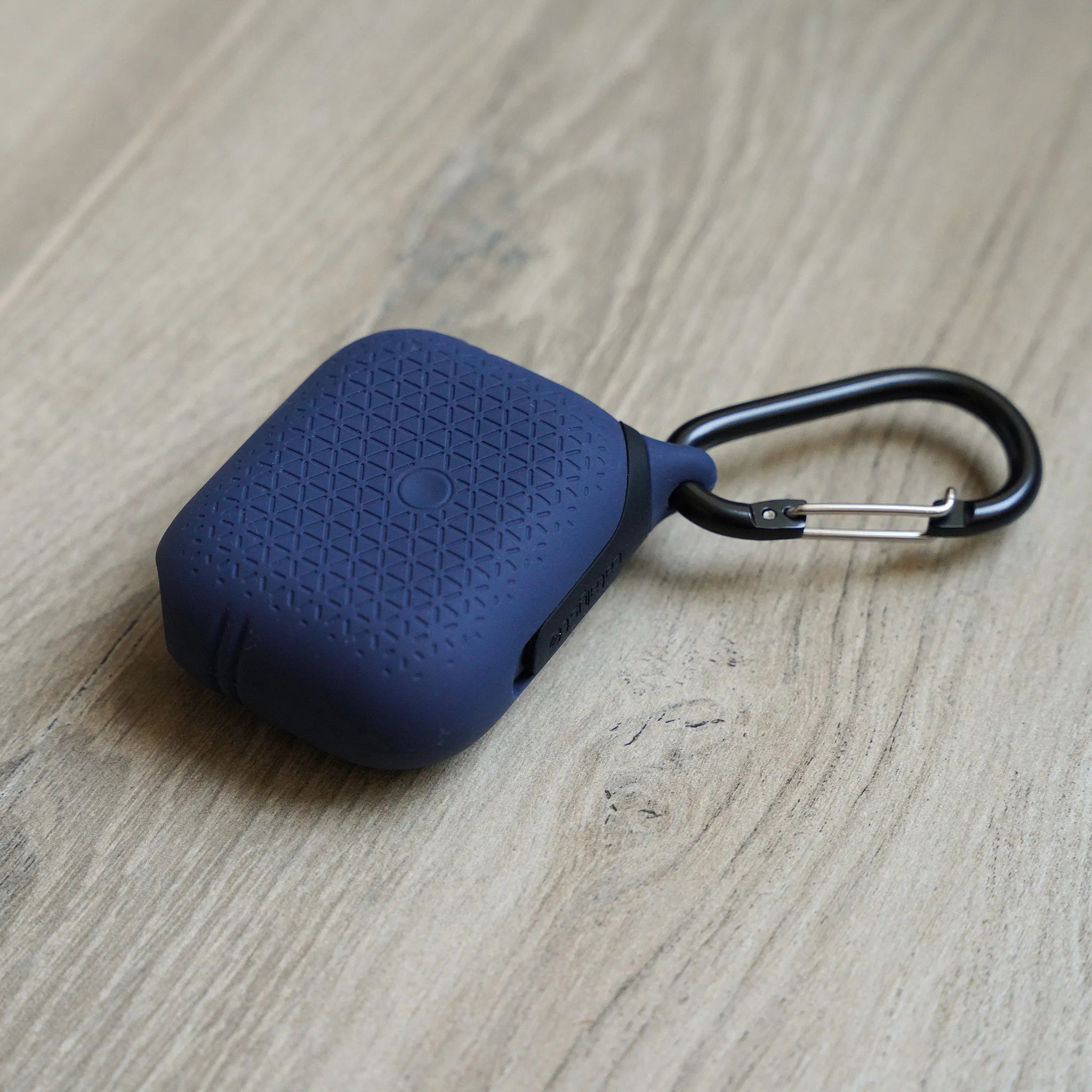 The Catalyst Case US AirPods Pro (Gen 2/1) Waterproof Case + Carabiner, in dark blue textured silicone with a circular pattern, securely holds the earbuds and sits diagonally on a light wood surface.