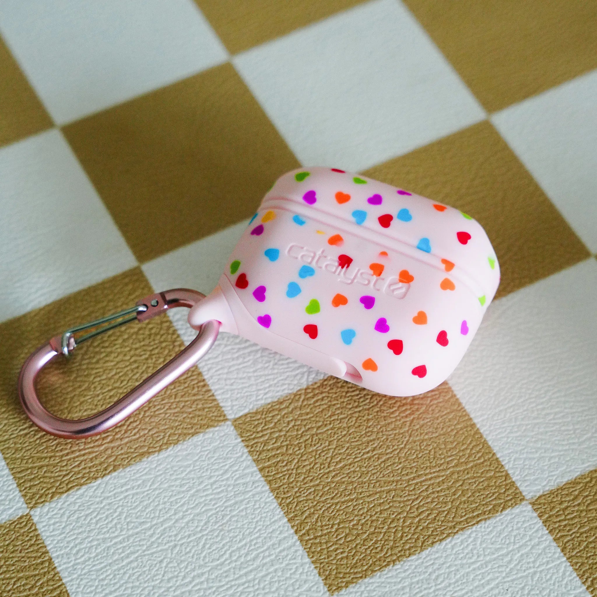 A pale pink Catalyst Case US AirPods Pro (Gen 2/1) Waterproof Case with colorful heart-shaped dots and a metallic pink carabiner sits on a tan-and-white checkerboard textured surface.