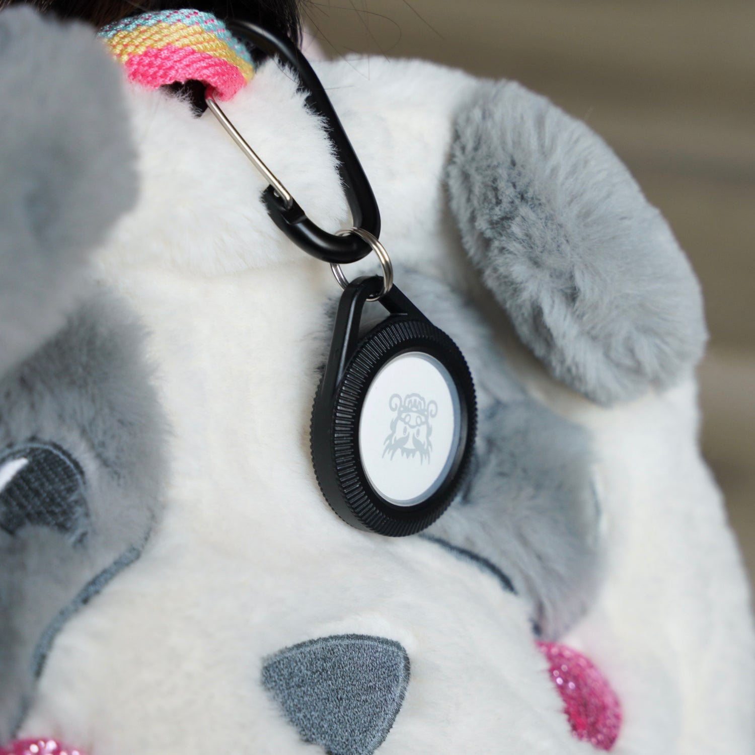 A close-up of a fluffy white and gray bag with animal features features the Catalyst Case US AirTag - Total Protection Case, Hang-It, attached by a black carabiner to a rainbow-striped strap for extra style and security.