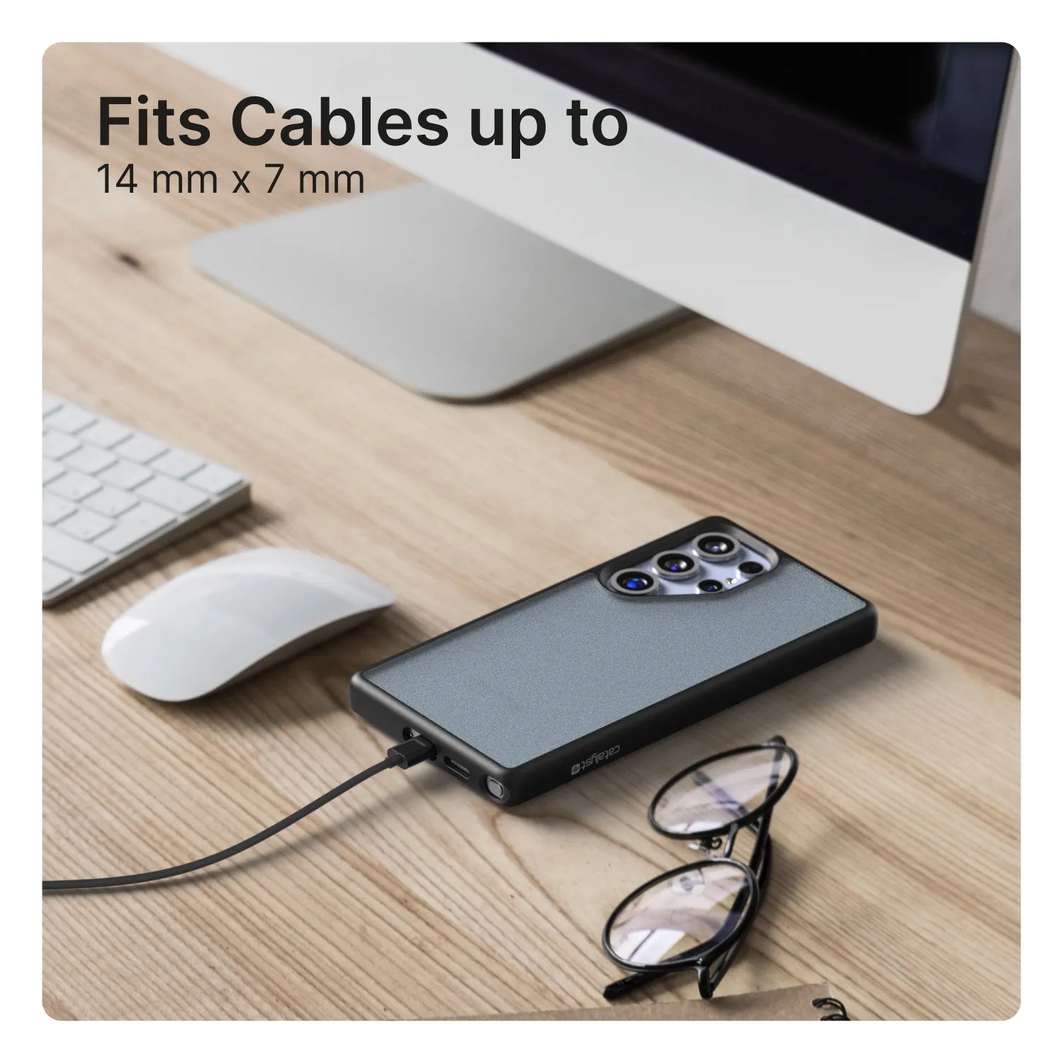 A Samsung Galaxy S25 Ultra in a Catalyst Case US Impact Protection Case (Pre-Order) rests on a wooden desk, charging via cable. Nearby are eyeglasses, mouse, keyboard, and monitor. Text reads: “Fits Cables up to 14 mm x 7 mm.” The setup is tidy and modern.