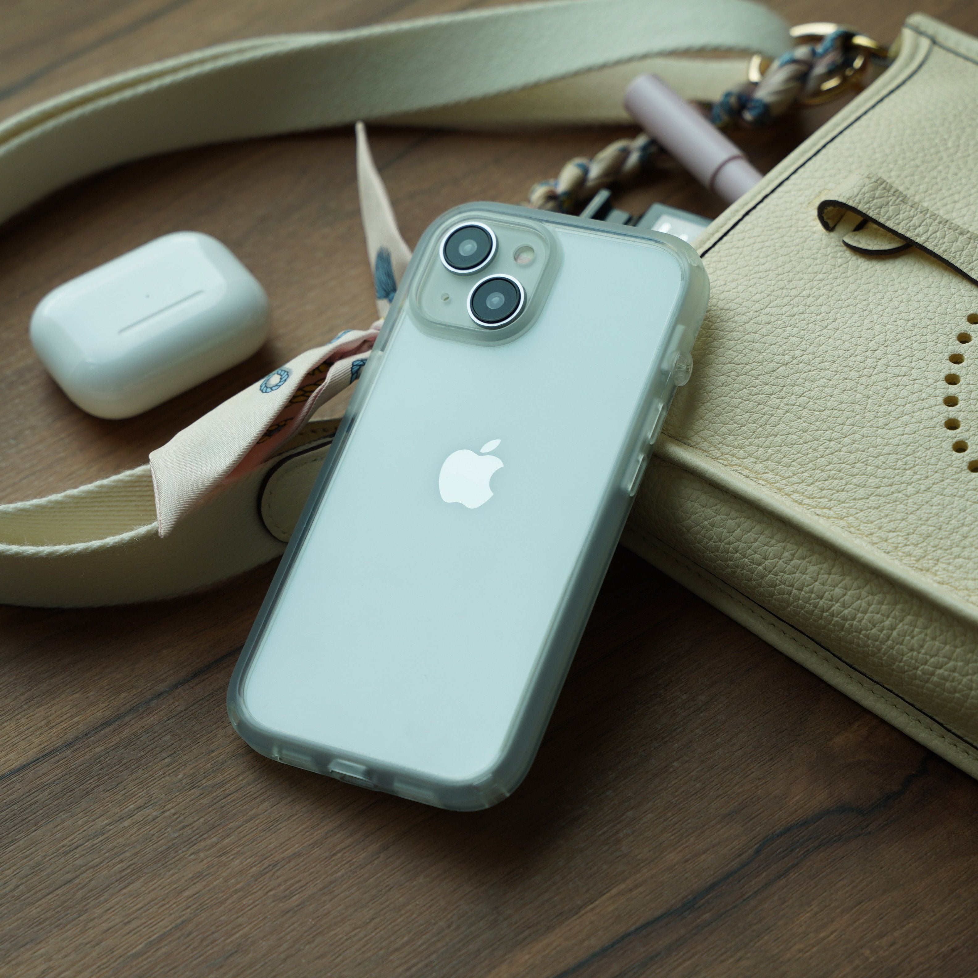 A Catalyst Case US iPhone 16 Plus with the clear Influence Case rests on a wooden surface beside a cream purse, scarf-detailed strap, and closed white AirPods case, creating a tidy and stylish scene.