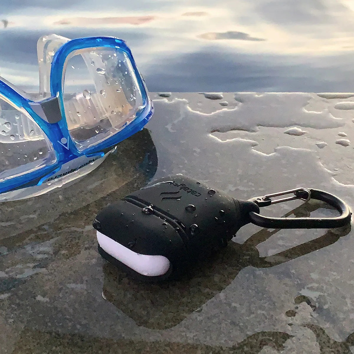 A Catalyst Case US AirPods (Gen 2/1) Waterproof Case with a carabiner rests on a wet stone surface, glistening with water droplets. The calm, reflective water and cloudy sky create a serene backdrop.