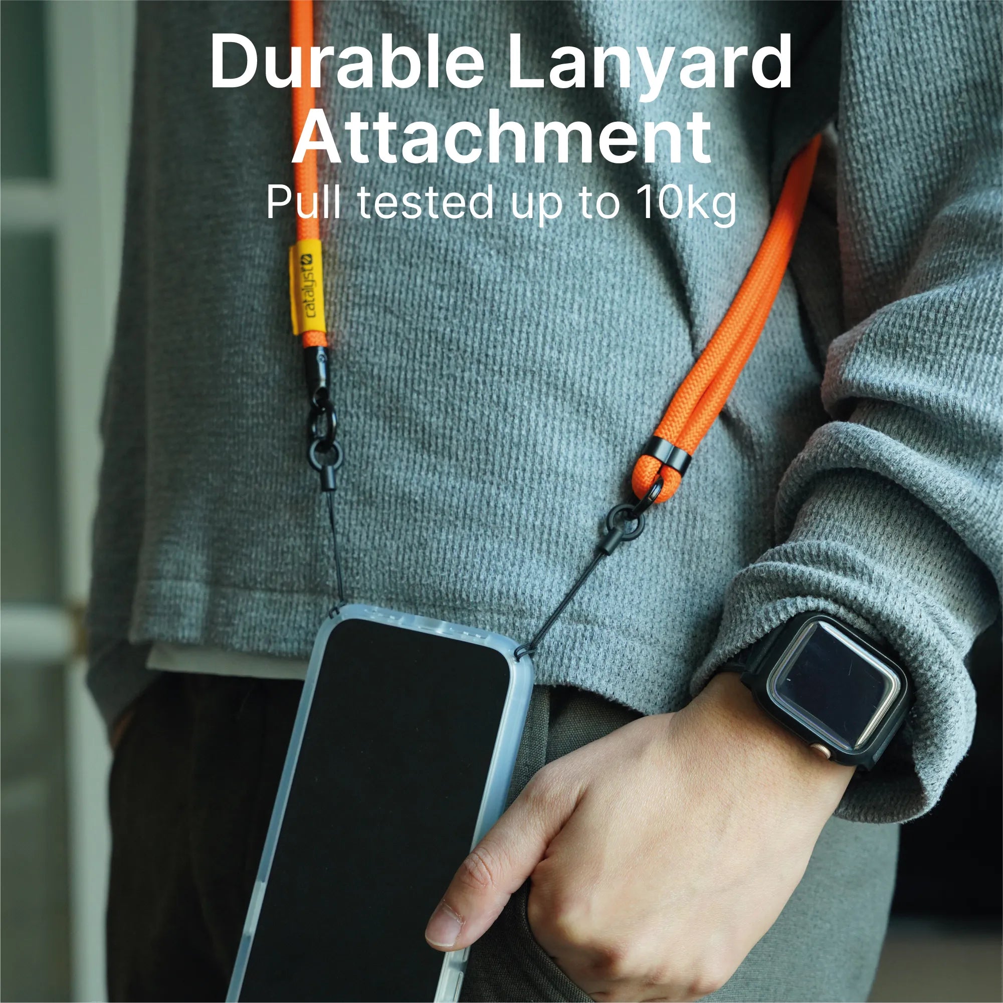 A person wearing a grey sweater and smartwatch holds a phone attached to an orange Catalyst Case US Crossbody Shoulder Strap. White text reads: "Durable Lanyard Attachment. Pull tested up to 10kg.