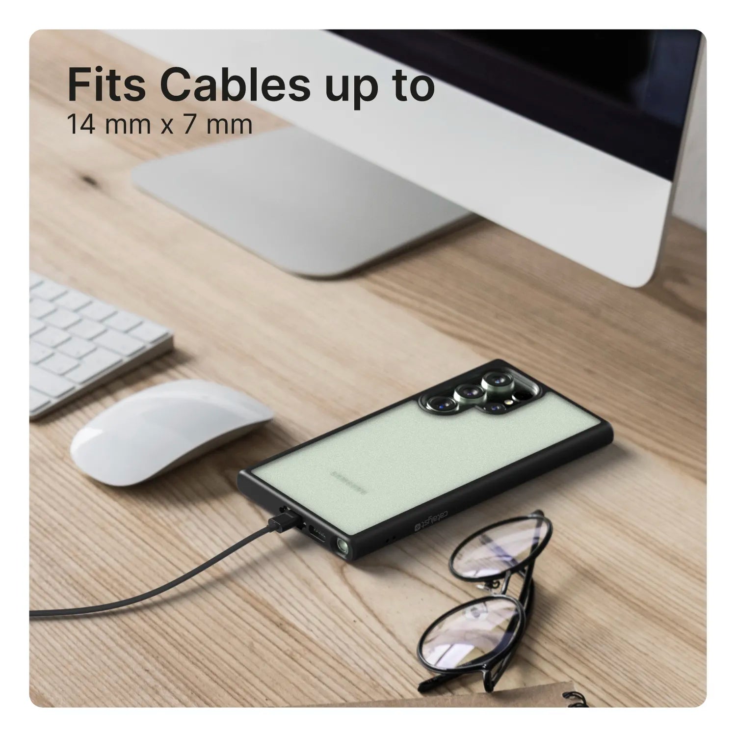 A Samsung Galaxy S23 Ultra with a Catalyst Case US Impact Protection Case charges on a wooden desk near an iMac, keyboard, mouse, and glasses. The image says “Fits Cables up to 14 mm x 7 mm,” highlighting the connected charging cable.