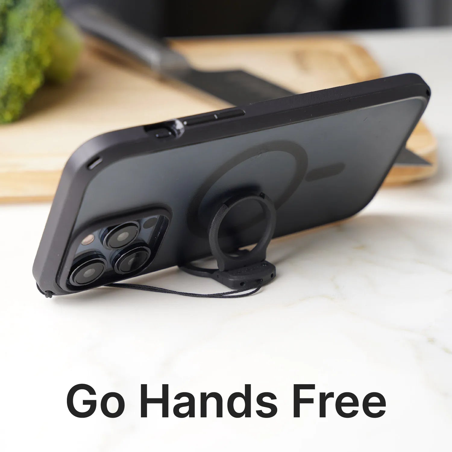 A Catalyst Case US smartphone with a Shoulder Strap & Ring Stand Bundle is propped horizontally on a white surface. A circular ring stand supports the phone, with “Go Hands Free.” in large text. A cutting board and broccoli appear in the background.
