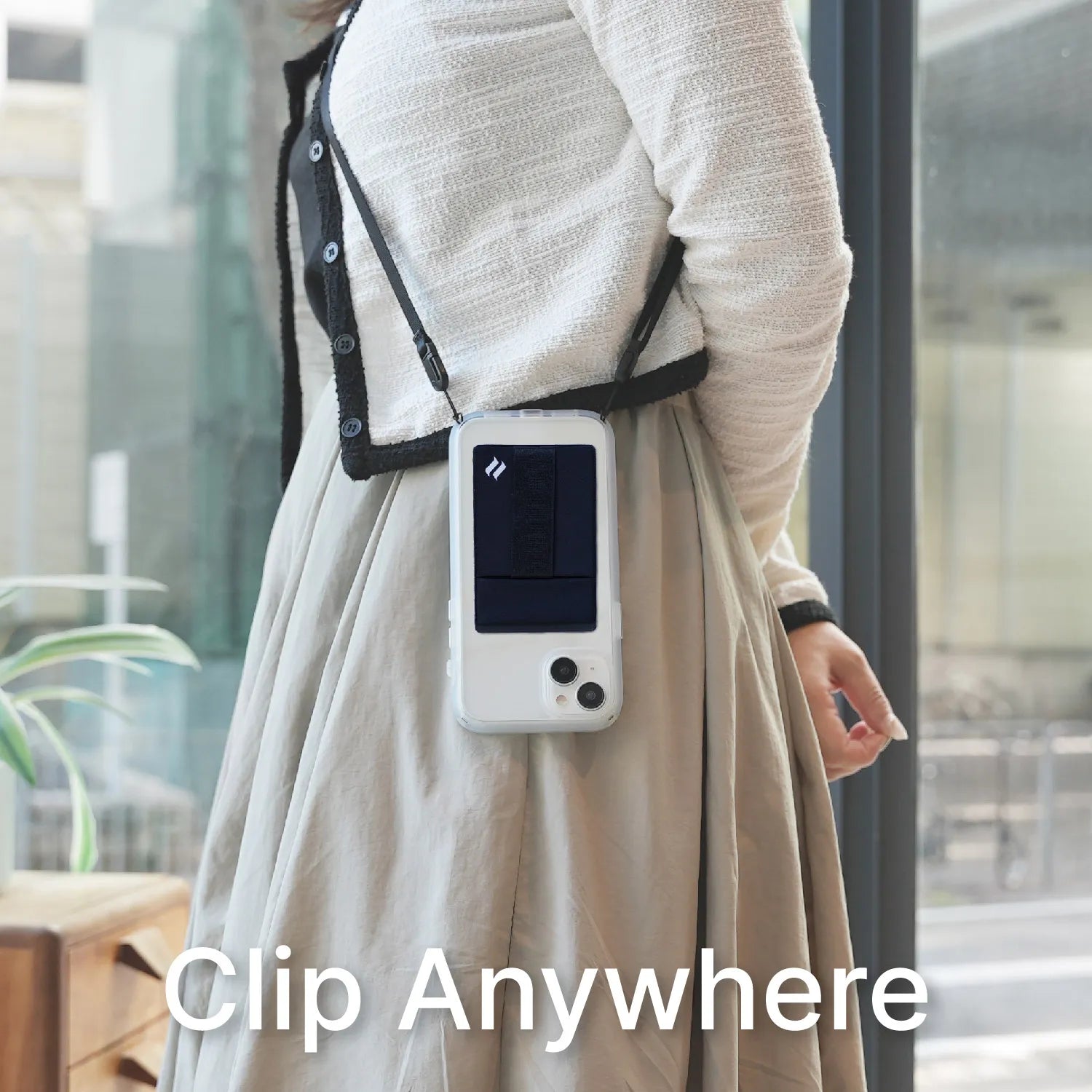 A woman wearing a light cardigan and beige skirt stands indoors with a smartphone in a Catalyst Case US Stick On Wallet & Shoulder Strap Bundle clipped to her waist.