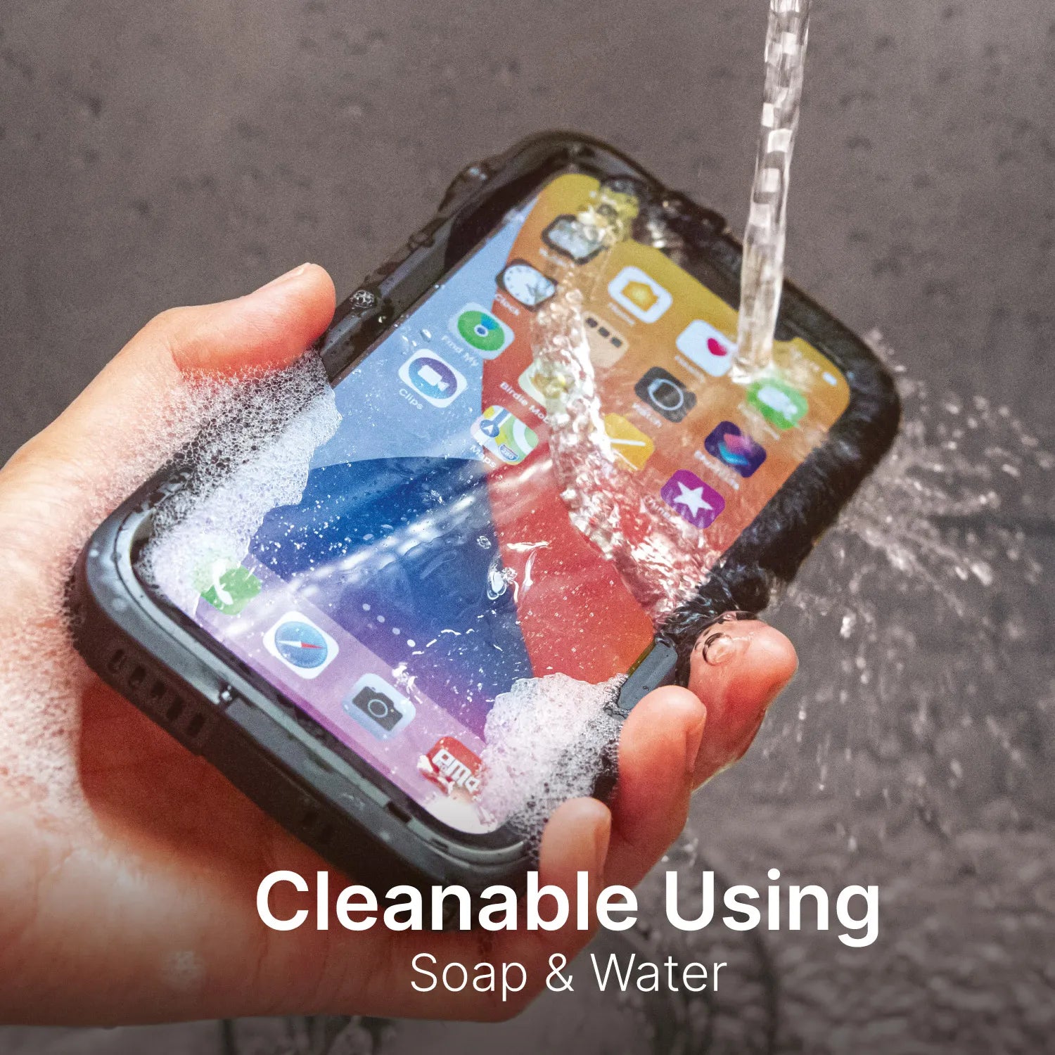 A hand holds an iPhone 12 Pro in a Catalyst Case US Waterproof Case under running water and soap suds. Colorful app icons appear on the screen. Bottom text: “Cleanable Using Soap & Water.” Total protection for your device.