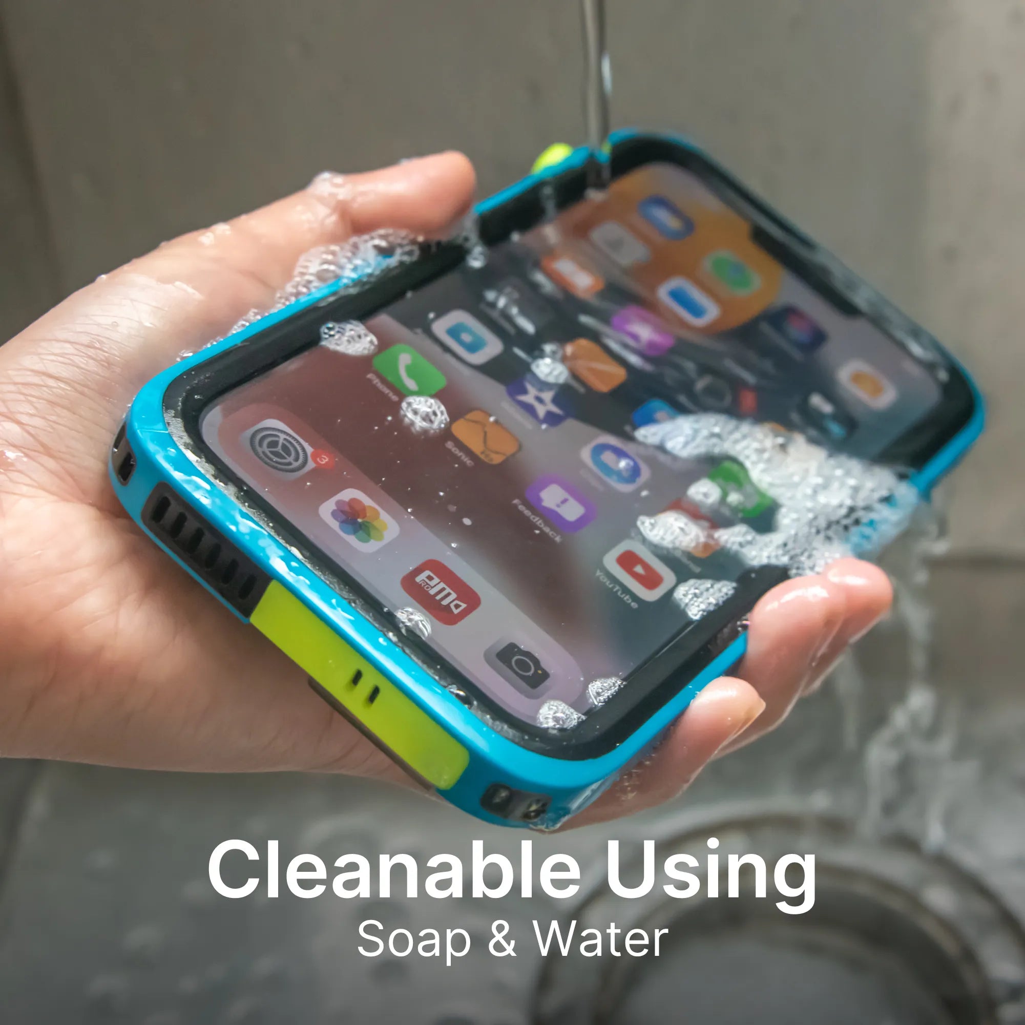A hand holds an iPhone 13 Pro Max in a blue and yellow Catalyst Case US waterproof case, under running water and soap suds. The phone’s screen shows app icons. Text below reads: “Cleanable Using Soap & Water.”.