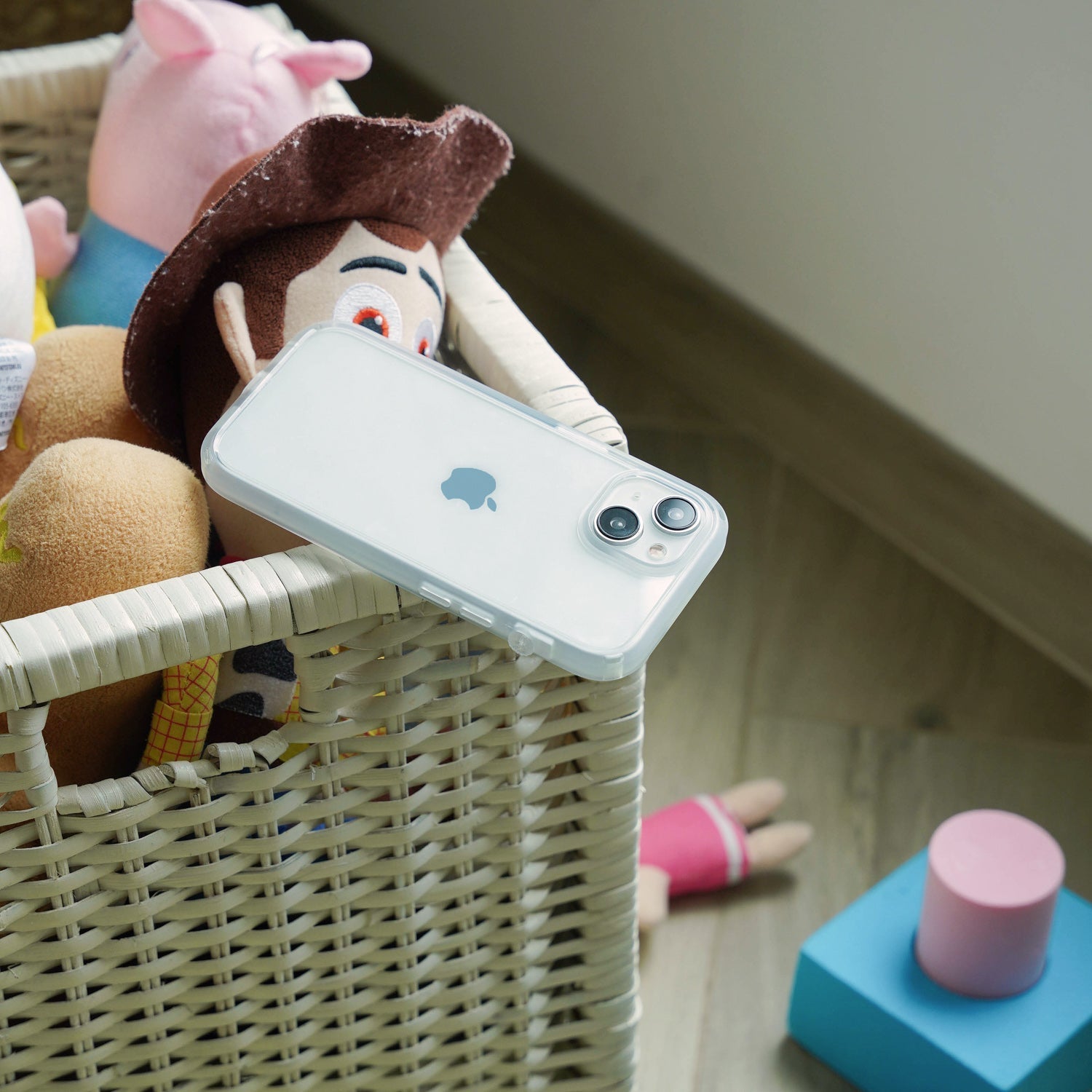 A wicker basket of plush toys, including a cowboy doll, sits on a wooden floor. An iPhone 14 in a Catalyst Case US Influence Case rests on the basket’s edge, with colorful blocks scattered nearby as sunlight streams in from the right.