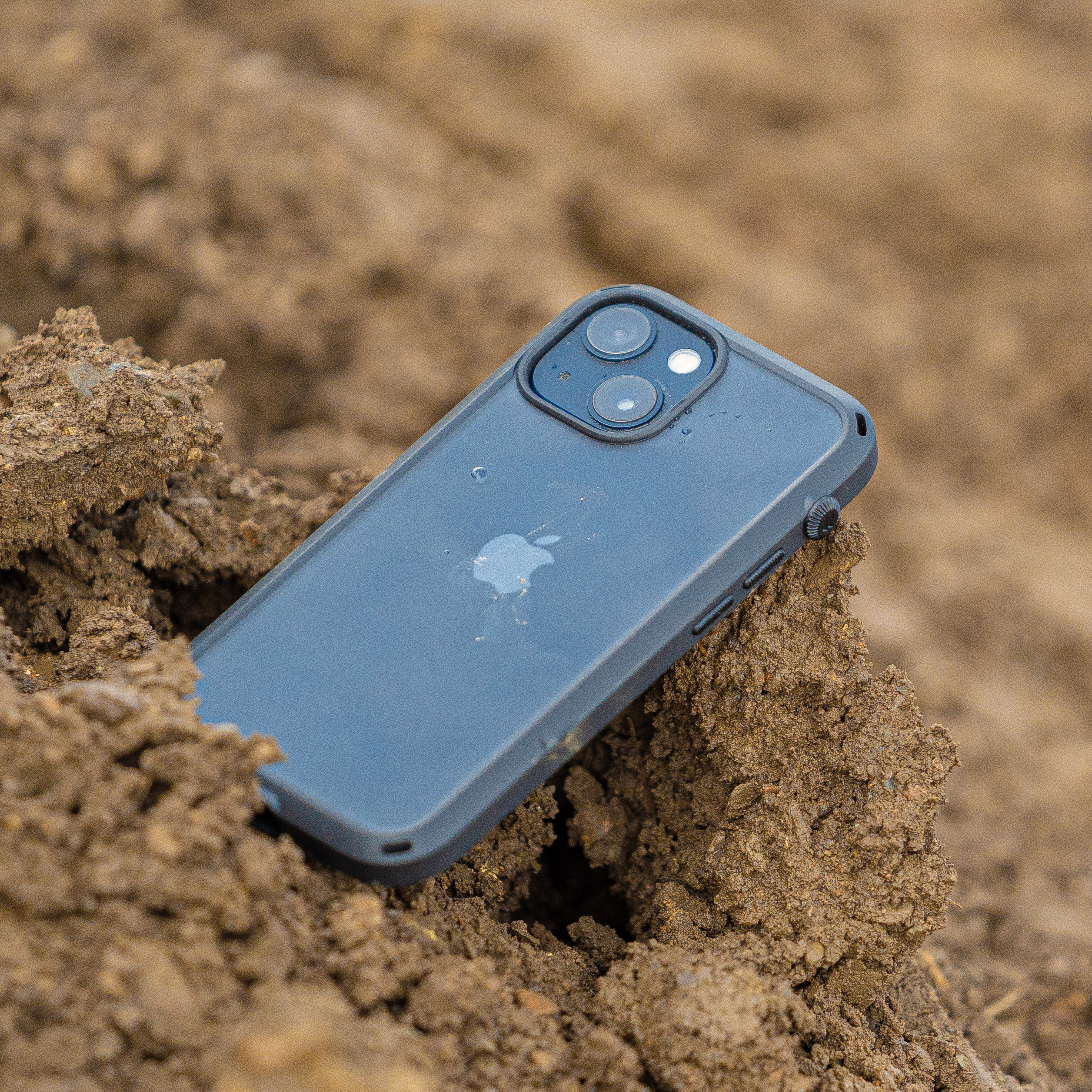 A Catalyst Case US iPhone 14 - Influence Case with a blue finish and visible Apple logo lies face down on rough brown dirt, partially buried. Dual camera lenses and rugged design hint at protection for outdoor adventures.