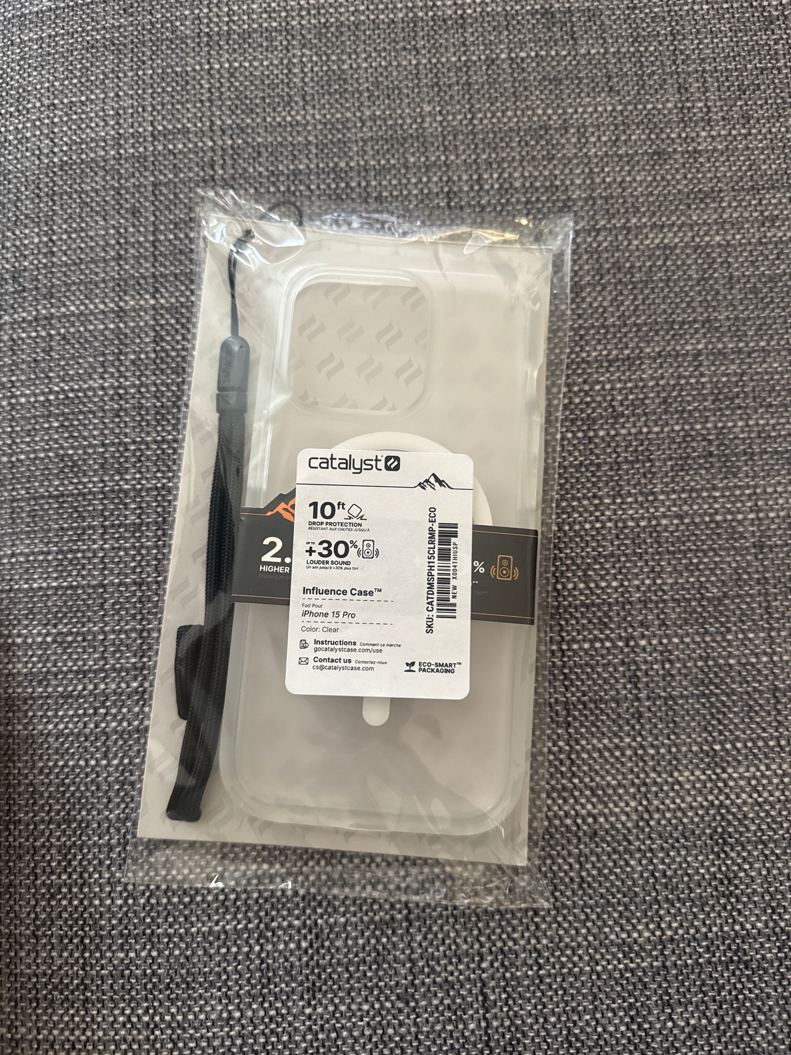 A packaged Catalyst Case US Influence Case for iPhone 15 Pro, MagSafe compatible, is displayed on a gray textured surface. The clear protective phone case is visible inside the plastic wrap and comes with a black lanyard attached. The label highlights features such as military-grade drop protection, dust proofing, and more.