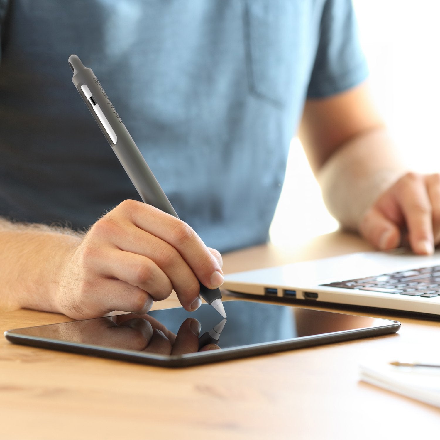 A person wearing a blue t-shirt uses the Apple Pencil (Gen 1) with a Catalyst Case US Grip Case on a tablet lying flat on a wooden desk, while their other hand rests on a laptop keyboard. The focus is on the stylus and tablet.