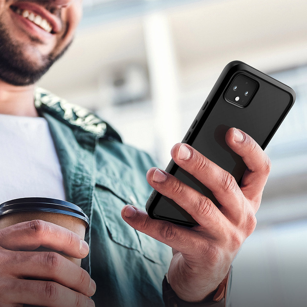 A smiling man holds a Google Pixel 4 XL with a Catalyst Case US Impact Protection Case in his right hand and a coffee cup in his left. He wears a green jacket over a white shirt, standing outdoors against a blurred urban background.