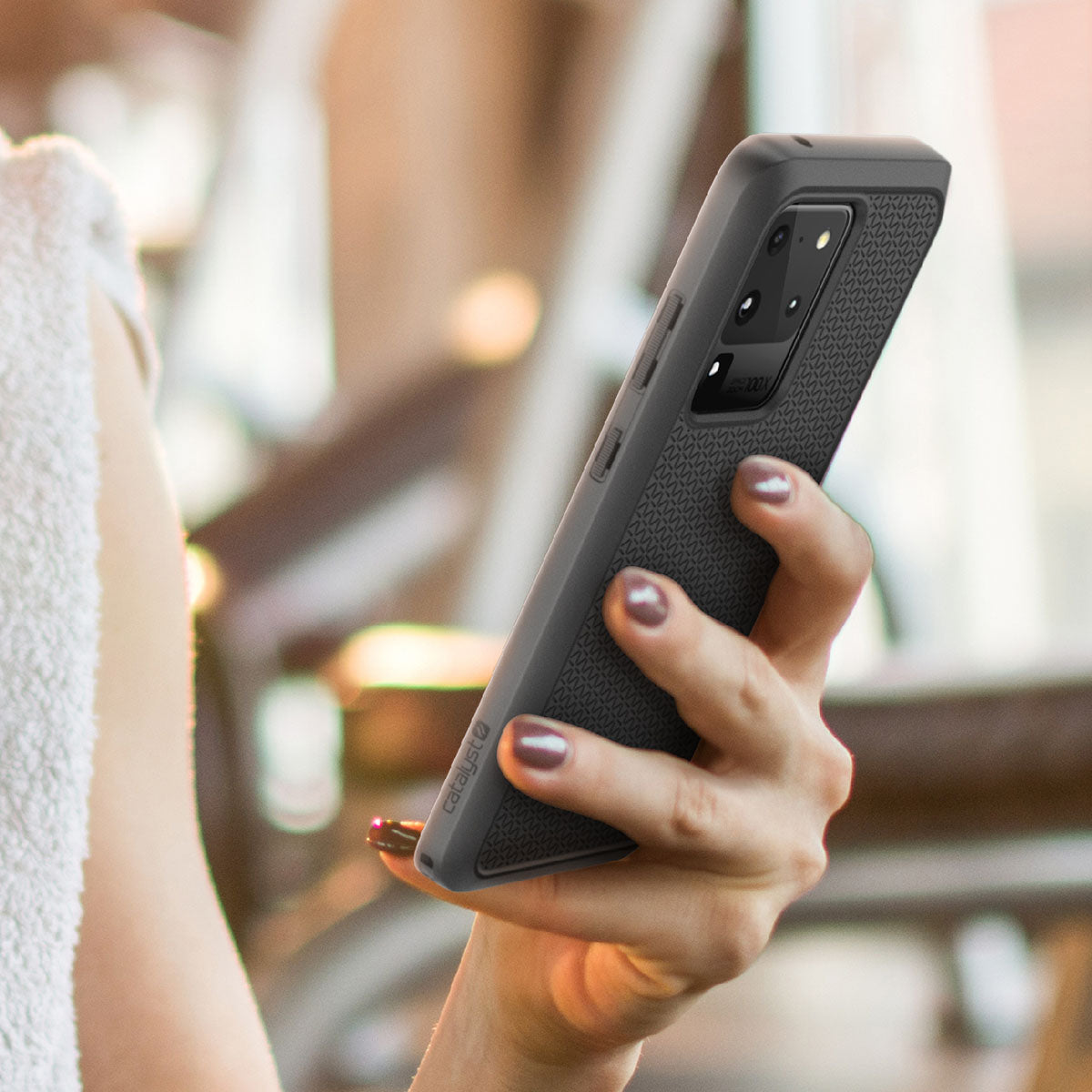 A person with manicured nails holds a Samsung Galaxy S20 Ultra in a textured dark Catalyst Case US Impact Protection Case. The phone’s camera module is visible, and part of a bare arm, white towel, and blurred gym equipment appear in the background.