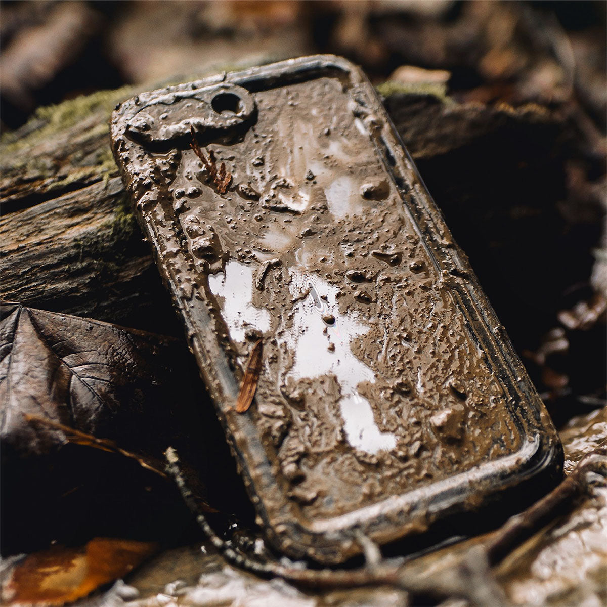 An iPhone 8 Plus in a Catalyst Case US Waterproof Case lies facedown in thick mud and forest debris, with its camera and rugged case barely visible among leaves and twigsâ€â€Âshowcasing its tough outdoor protection.