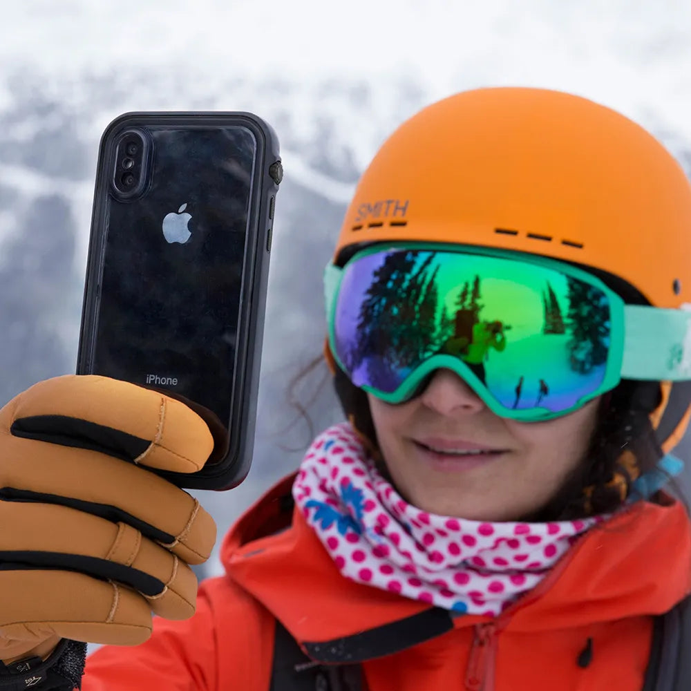 Wearing an orange helmet, mirrored ski goggles, and winter gear, a person holds up an iPhone Xs Max protected by a Catalyst Case US Waterproof Case in a snowy mountain setting. The goggles reflect snow-covered trees and sky.