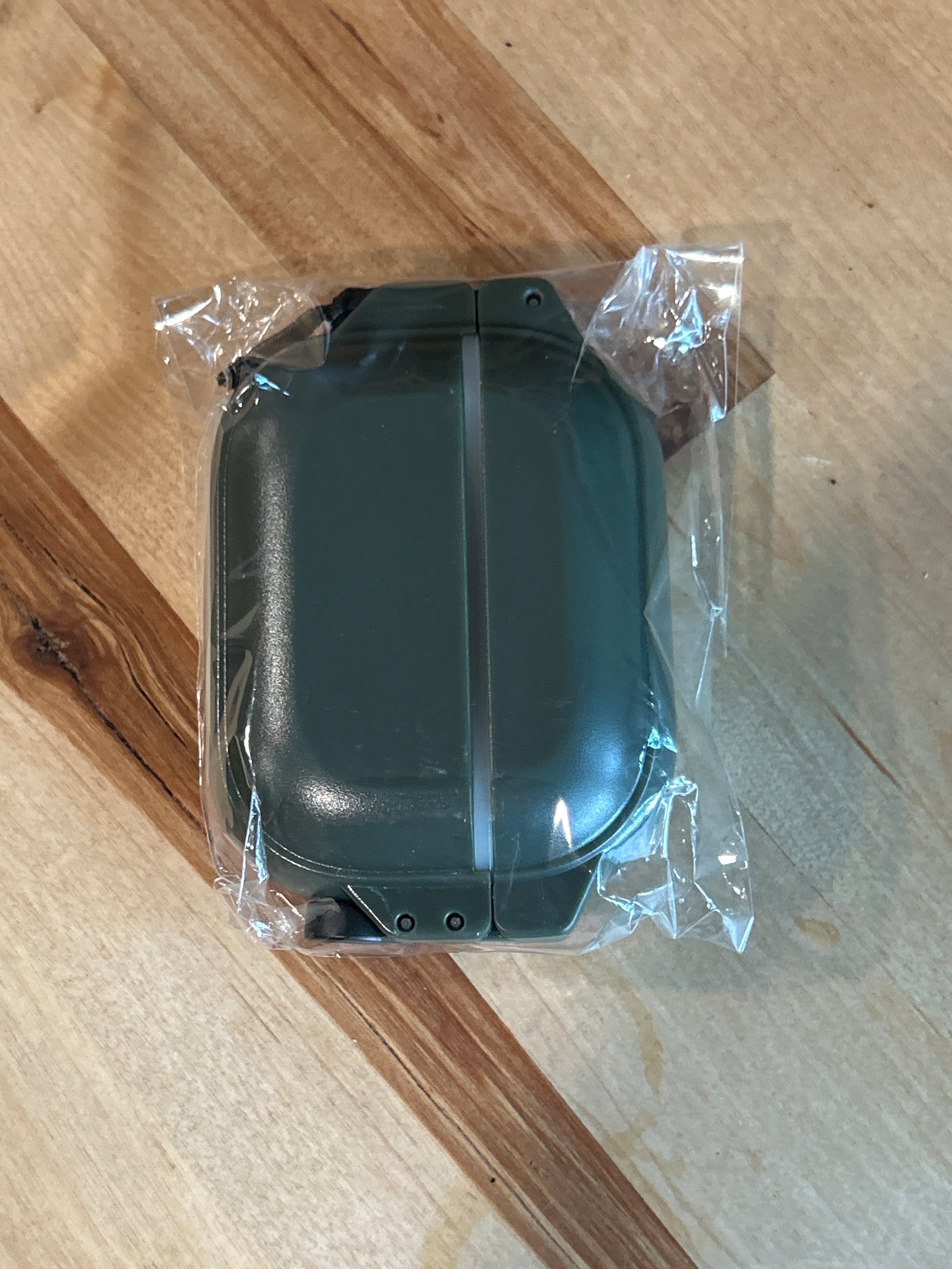 A green glow-in-the-dark rectangular plastic case, the AirPods Pro 3 - 100M Waterproof Total Protection Case + Carabiner by Catalyst Case US, featuring military-grade protection, is securely sealed in a clear plastic wrapper and rests on a wooden surface with visible grain and knots. The case appears to be new and unused.