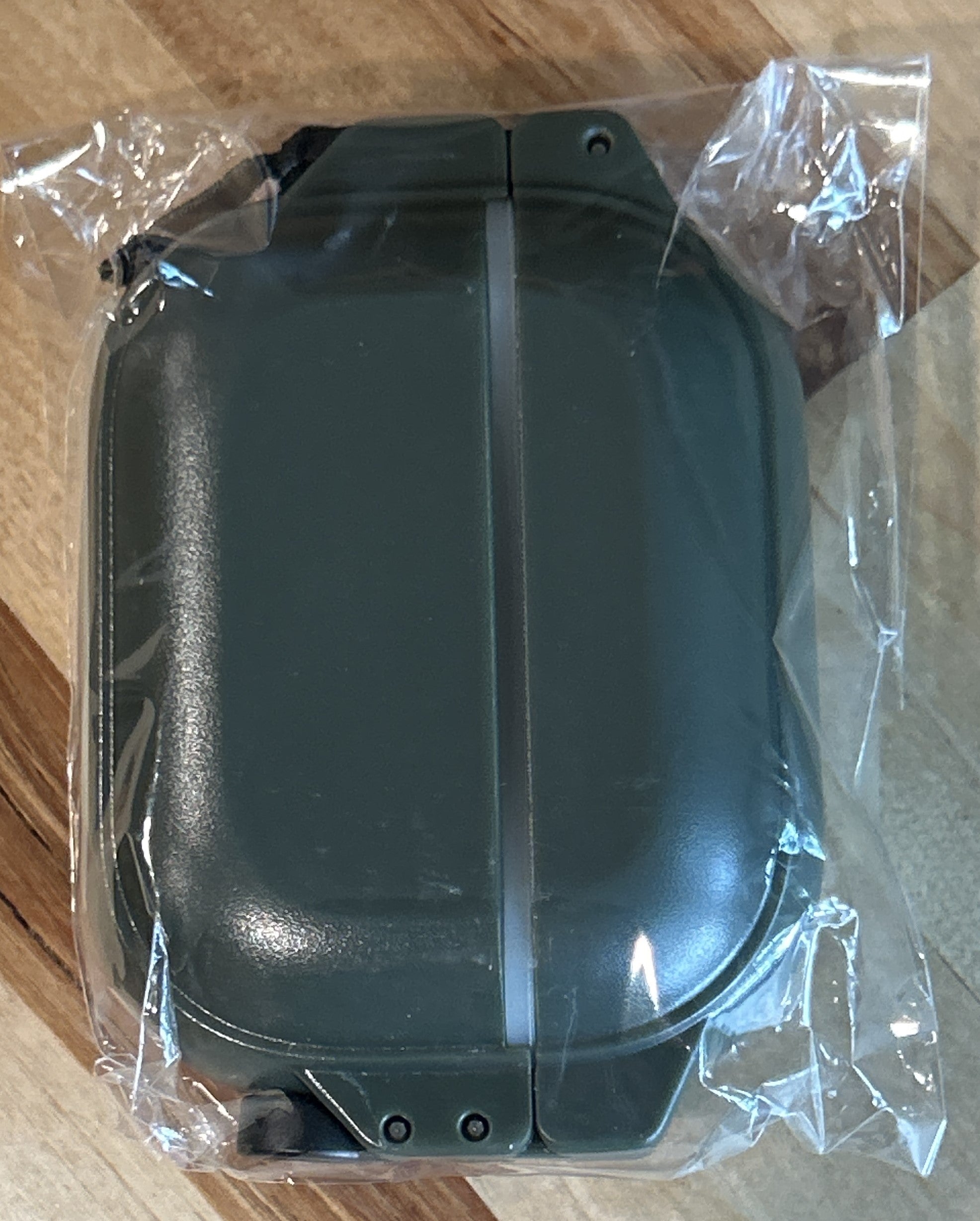 A compact, rectangular Catalyst Case US AirPods Pro 3 – 100M Waterproof Total Protection Case + Carabiner in a glow-in-the-dark green finish features rounded edges and military-grade drop protection. It is sealed in clear, crinkled plastic packaging, closed and resting on a wooden surface with visible grain patterns.