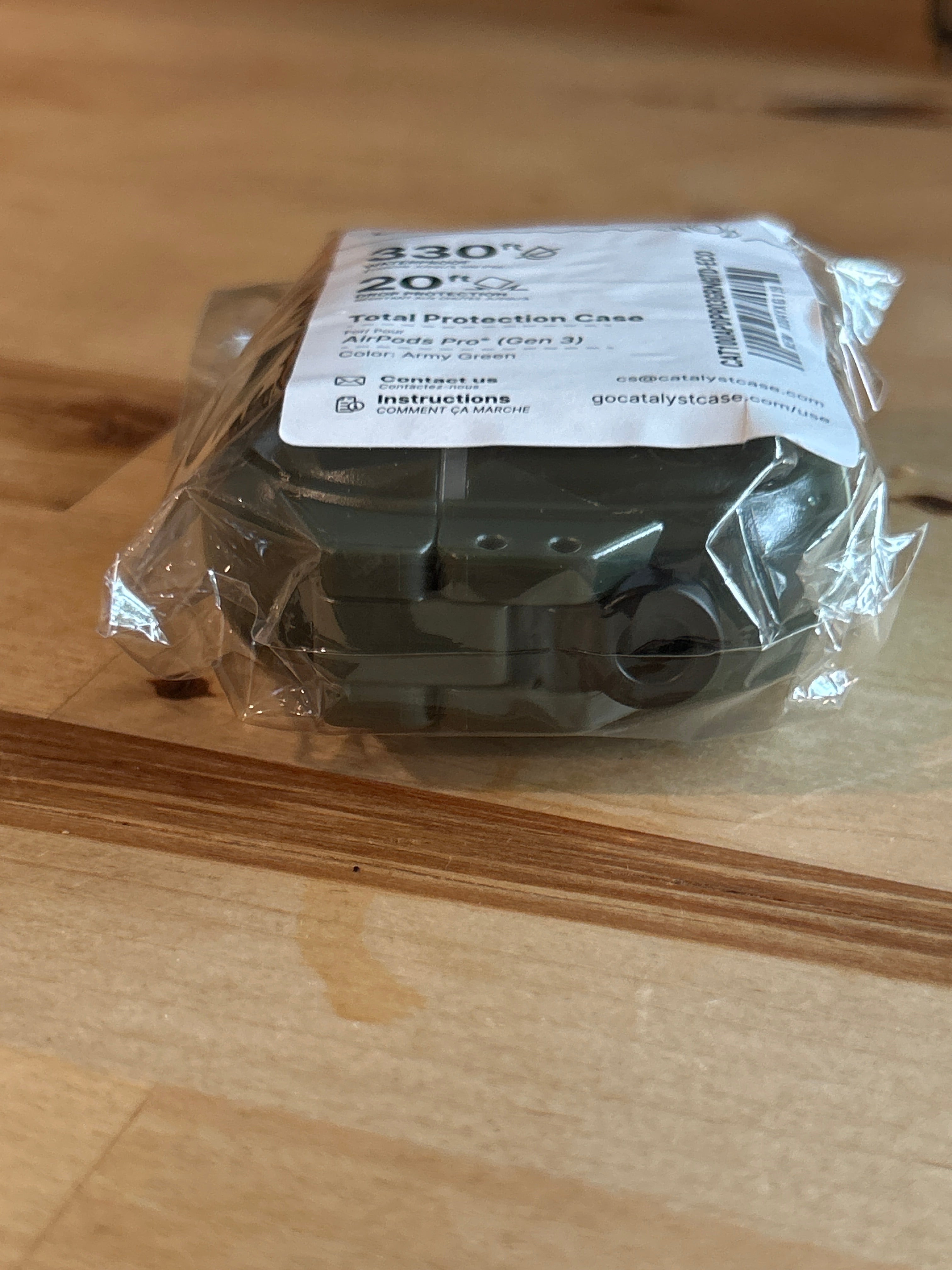 A green glow-in-the-dark Catalyst Case US “AirPods Pro 3 - 100M Waterproof Total Protection Case + Carabiner” is wrapped in clear plastic on a wooden table, with a white label on top displaying product details, pricing, and a barcode. The durable design suggests military-grade protection for AirPods Pro 3, while the wood grain is visible in the background.