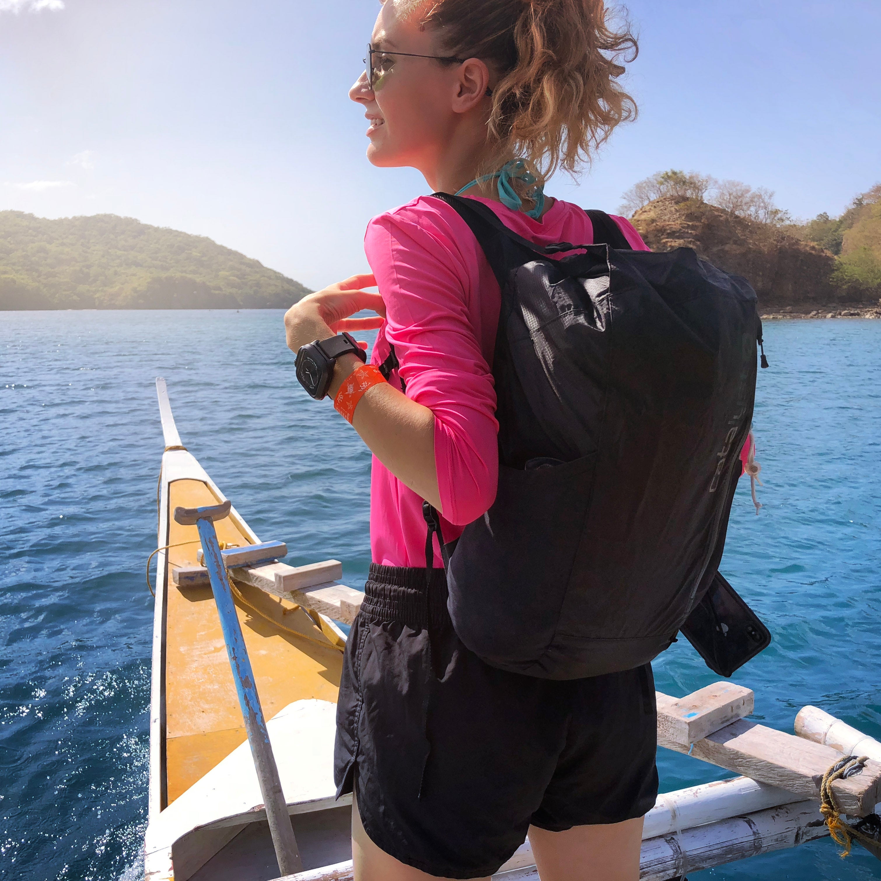 A person in a pink long-sleeve shirt and shorts stands on a boat, wearing a black Catalyst 20L Waterproof Backpack. It's a sunny vacation day with blue water and distant hills in the background.