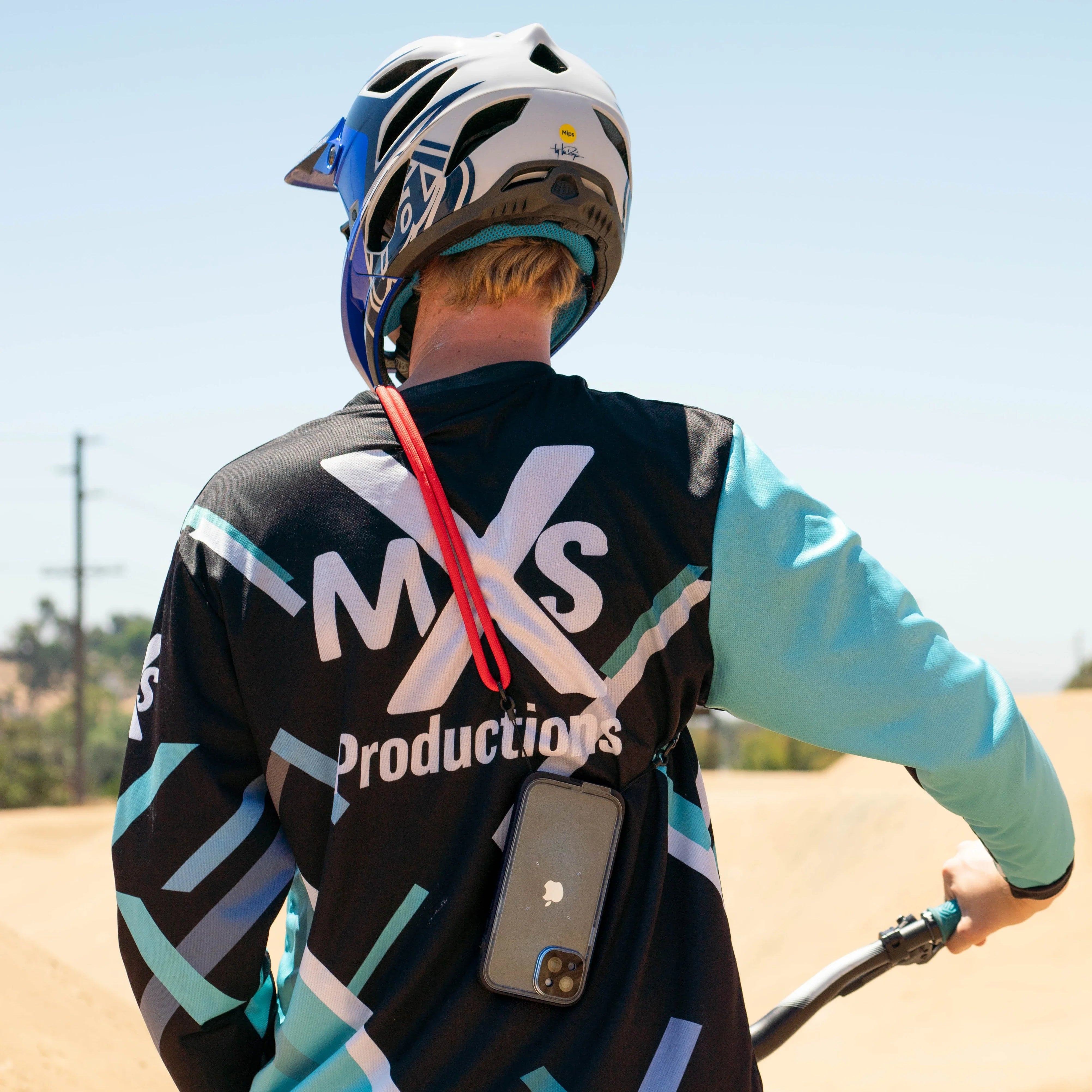 A person wearing a blue and black "MXS Productions" jersey and a helmet stands at a dirt bike track, with their back visible. A Catalyst Case US Crossbody Shoulder Strap holds their smartphone hands-free on the jersey.
