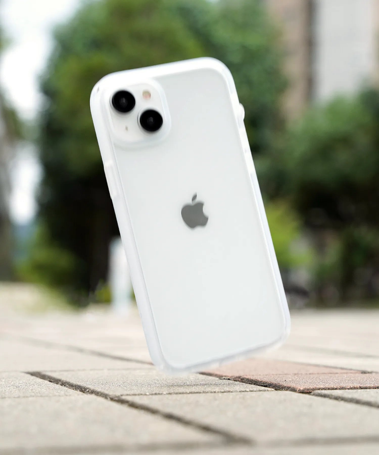 An iPhone with a clear Catalyst Drop Proof Case is shown mid-air falling on the tiles in the corner. The background is faded, and trees and greenery can be seen. 