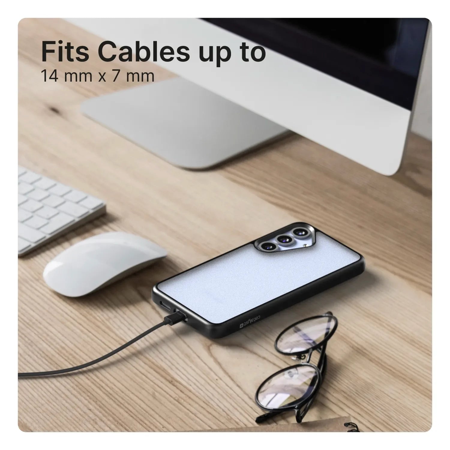 A Samsung Galaxy S25 Plus in a Catalyst Case US Impact Protection Case (Pre-Order) is connected to a charging cable on a tidy wooden desk near an iMac, keyboard, mouse, and eyeglasses. Text reads, "Fits Cables up to 14 mm x 7 mm.