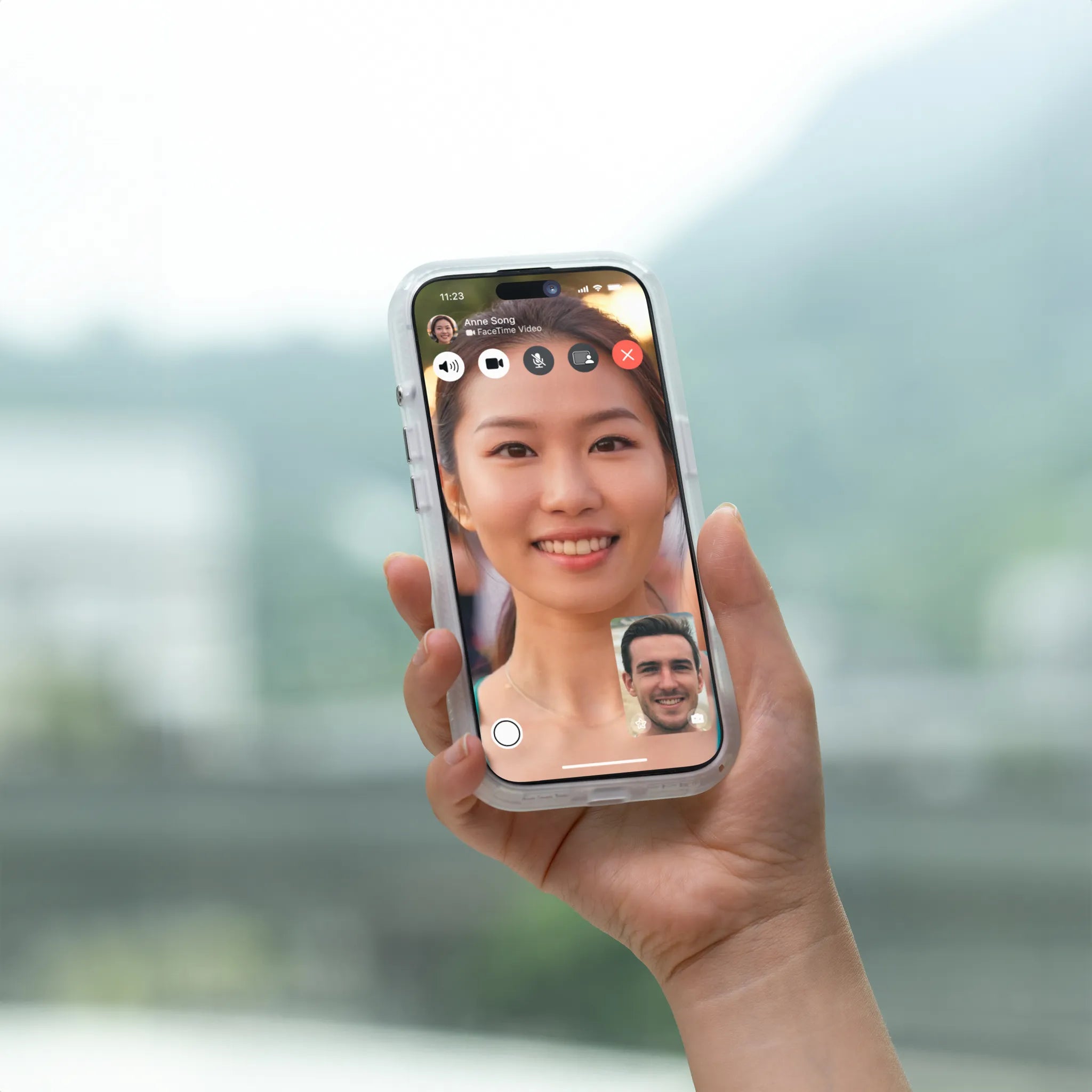 A hand holds an iPhone 15 Pro in a Catalyst Case US Influence Case (MagSafe Compatible), showing a video call with a smiling woman on the screen, a man in an inset, and blurred outdoor scenery with greenery and mountains in the background.