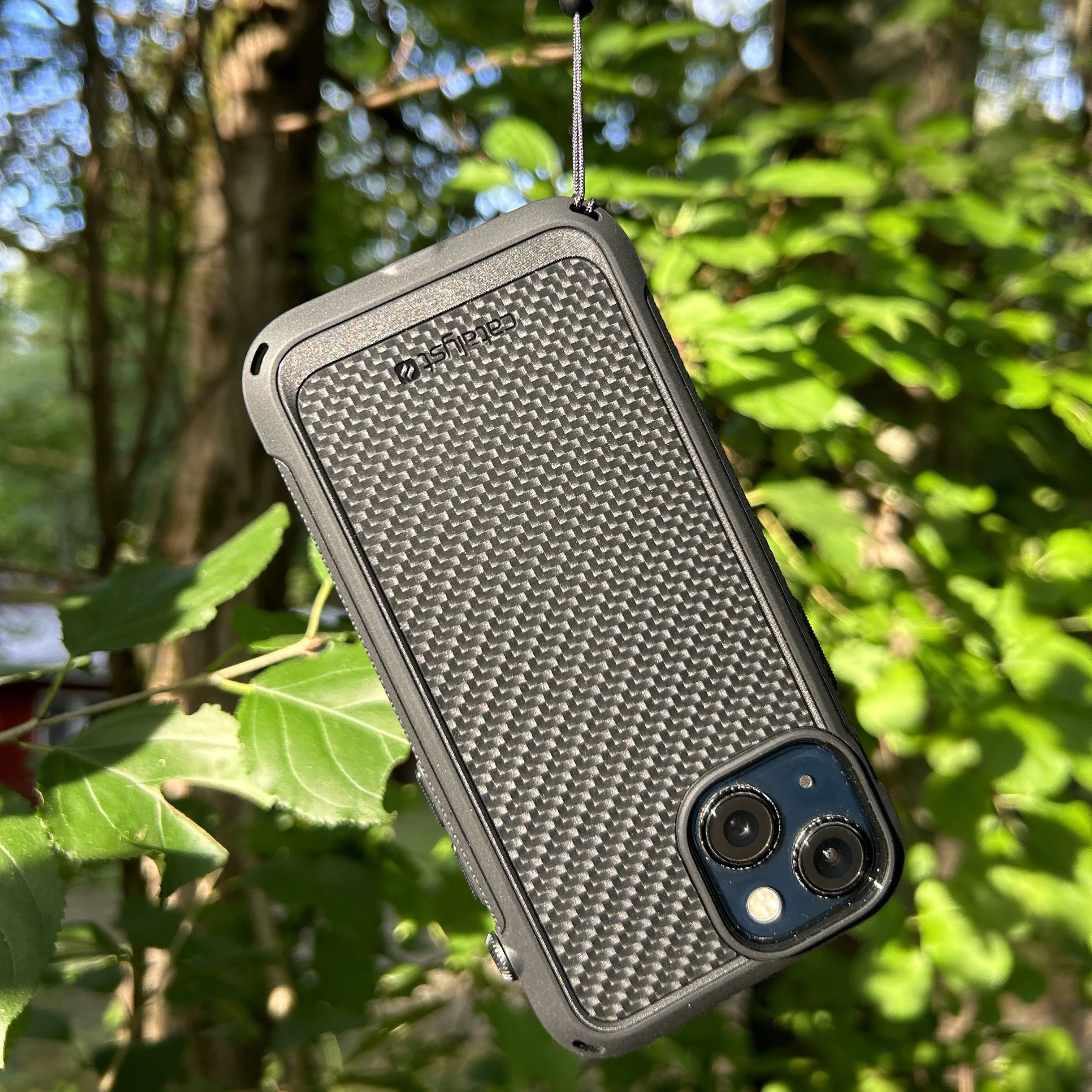 The Catalyst Case US iPhone 13 Pro Max Vibe Case (MagSafe Compatible) in textured black hangs by a black cord from a branch. Sunlight highlights the phone and green leaves, with blurred trees and foliage in the background.