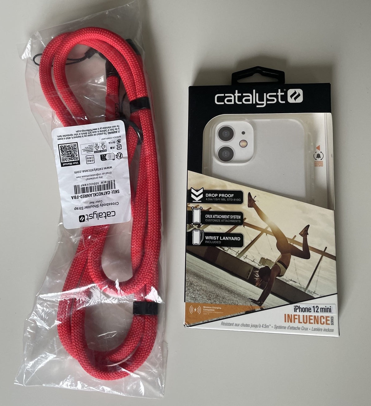 Side-by-side view of a packaged red Catalyst wrist lanyard next to the retail box for the Catalyst Influence Case for the iPhone 12 mini, a clear protective phone case engineered with military-grade drop protection.