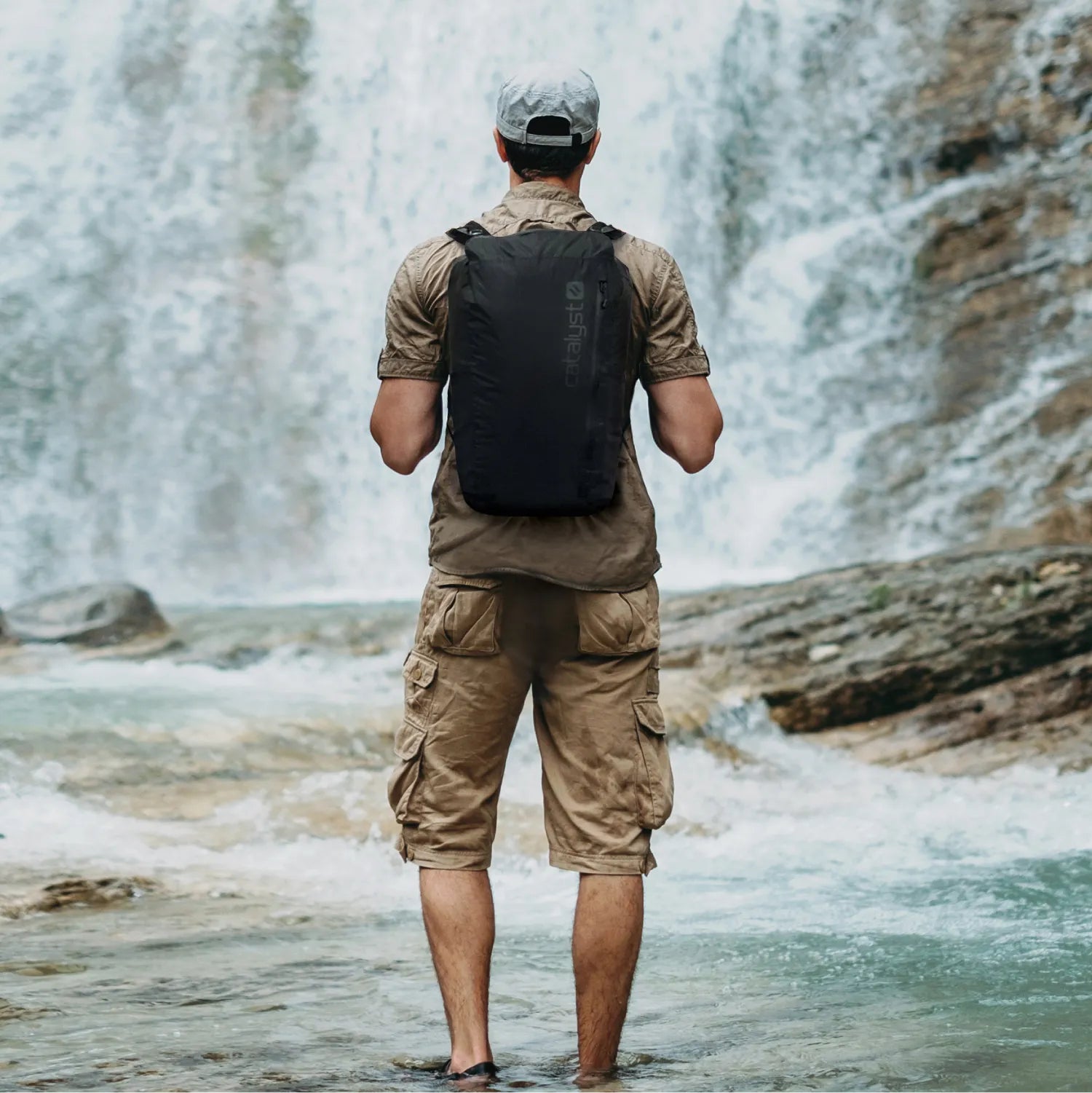Wearing a khaki shirt, cargo shorts, and a light gray cap, a person stands ankle-deep in water facing a waterfall. They carry the Catalyst Case US Waterproof 20L Backpack, surrounded by rocks and flowing water.
