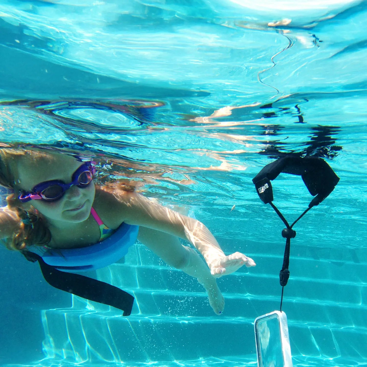 A young girl in purple goggles and a blue float swims underwater toward a Catalyst Case US Floating Lanyard - Stealth Black, which holds a phone case and floats nearby as sunlight reflects off the pool’s clear blue tiles.