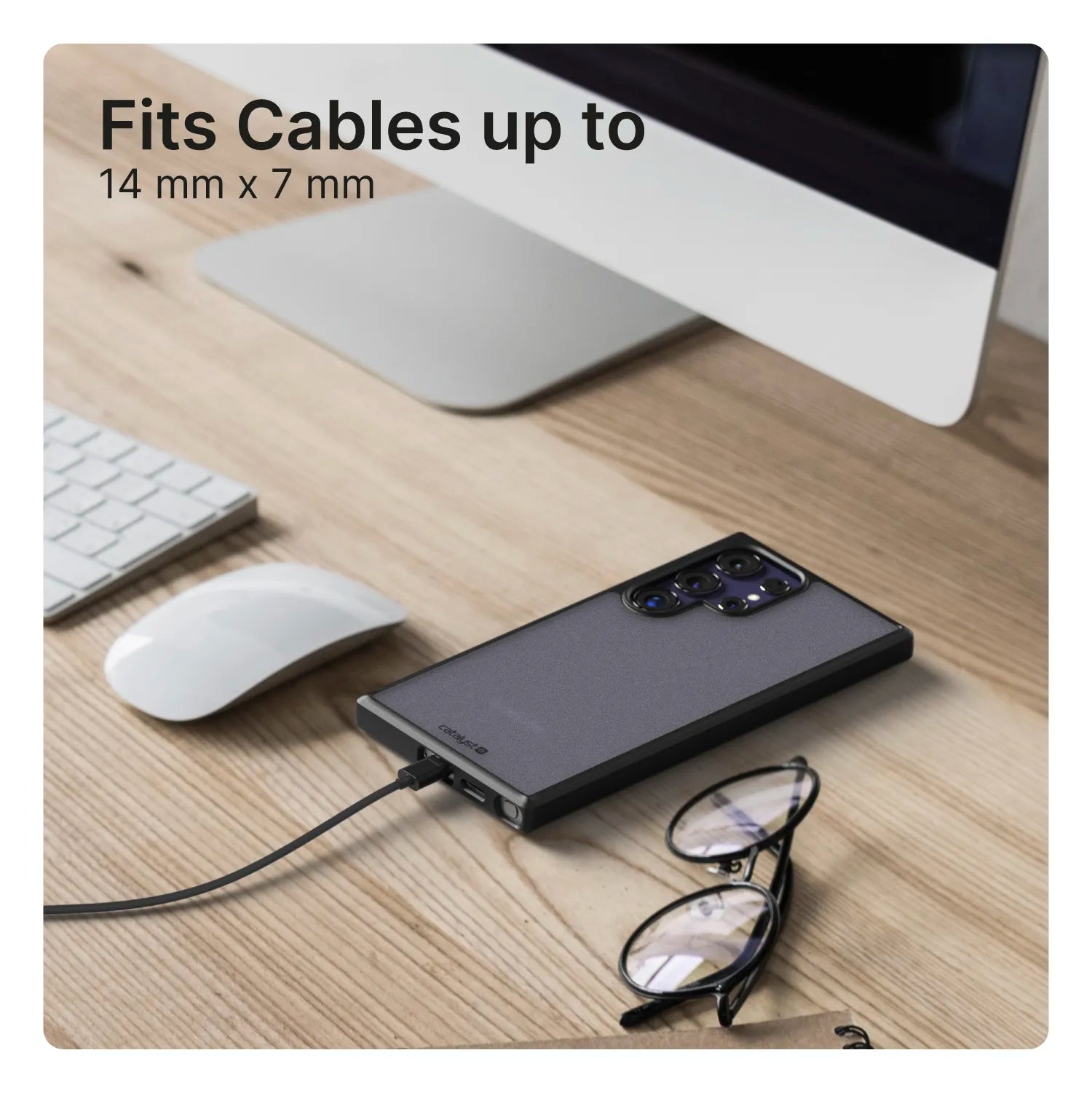A Samsung Galaxy S24 Ultra in a Catalyst Case US Impact Protection Case charges on a wooden desk beside a keyboard, monitor, and eyeglasses. Text in the top left reads "Fits Cables up to 14 mm x 7 mm.
