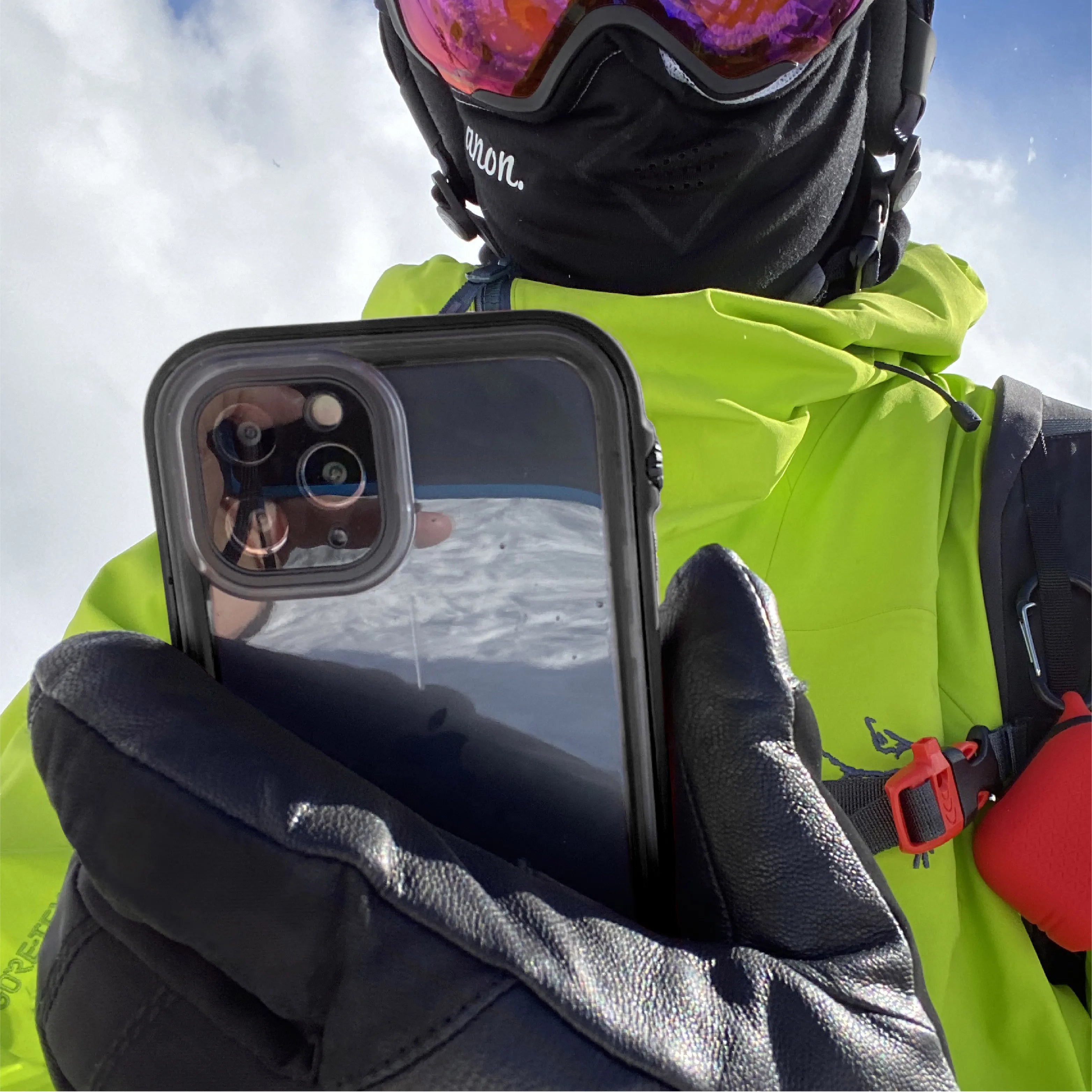 Wearing a neon green jacket, black gloves, and ski goggles, a person takes a selfie outdoors in snowy mountains using an iPhone 11 Pro Max with a Catalyst Case US waterproof case; snow and sky reflect in the phone.
