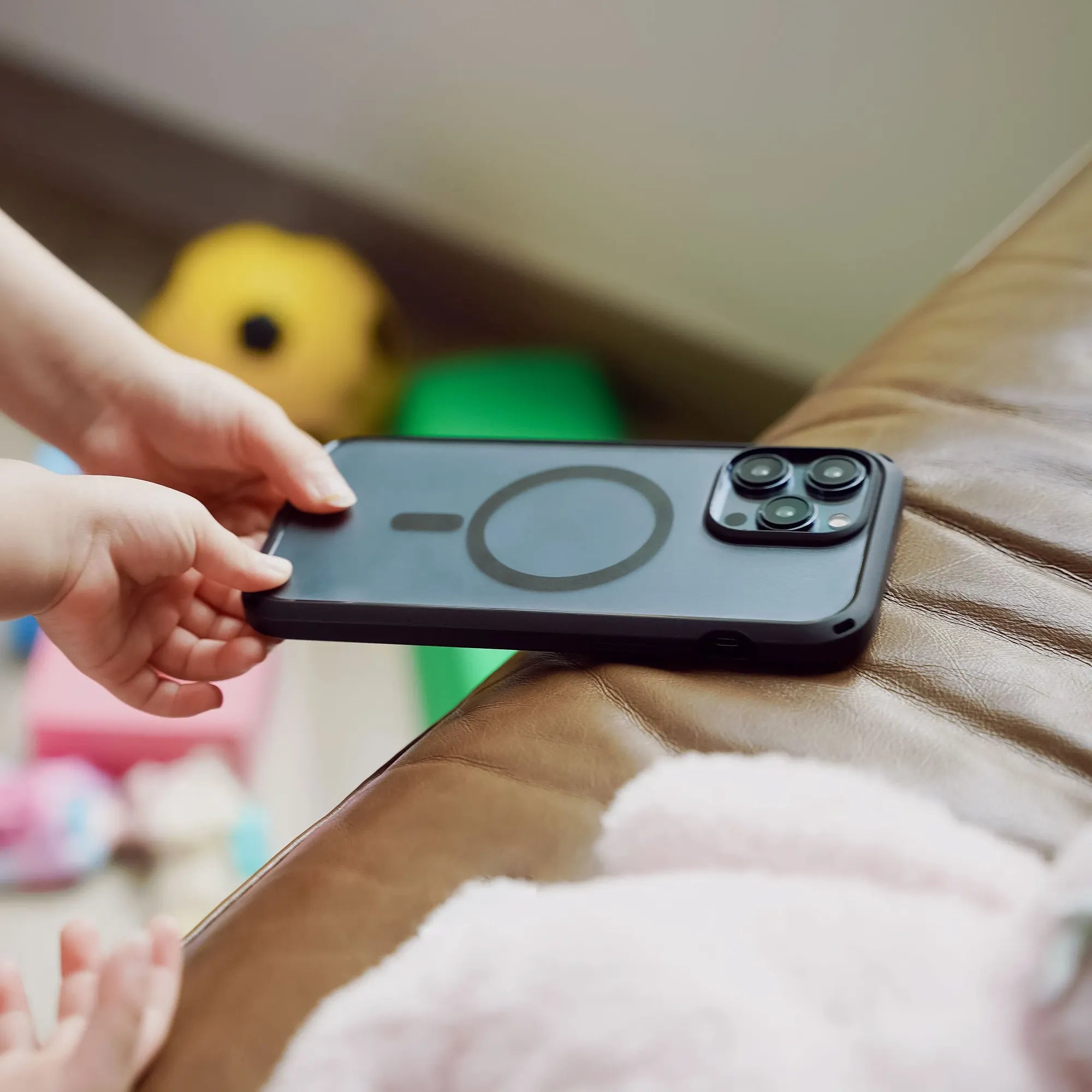 A child’s hands reach for a Catalyst Case US iPhone 14 Pro Max in an Influence Case (MagSafe Compatible) resting on a brown leather couch, with colorful toys and a plush animal scattered playfully in the background.