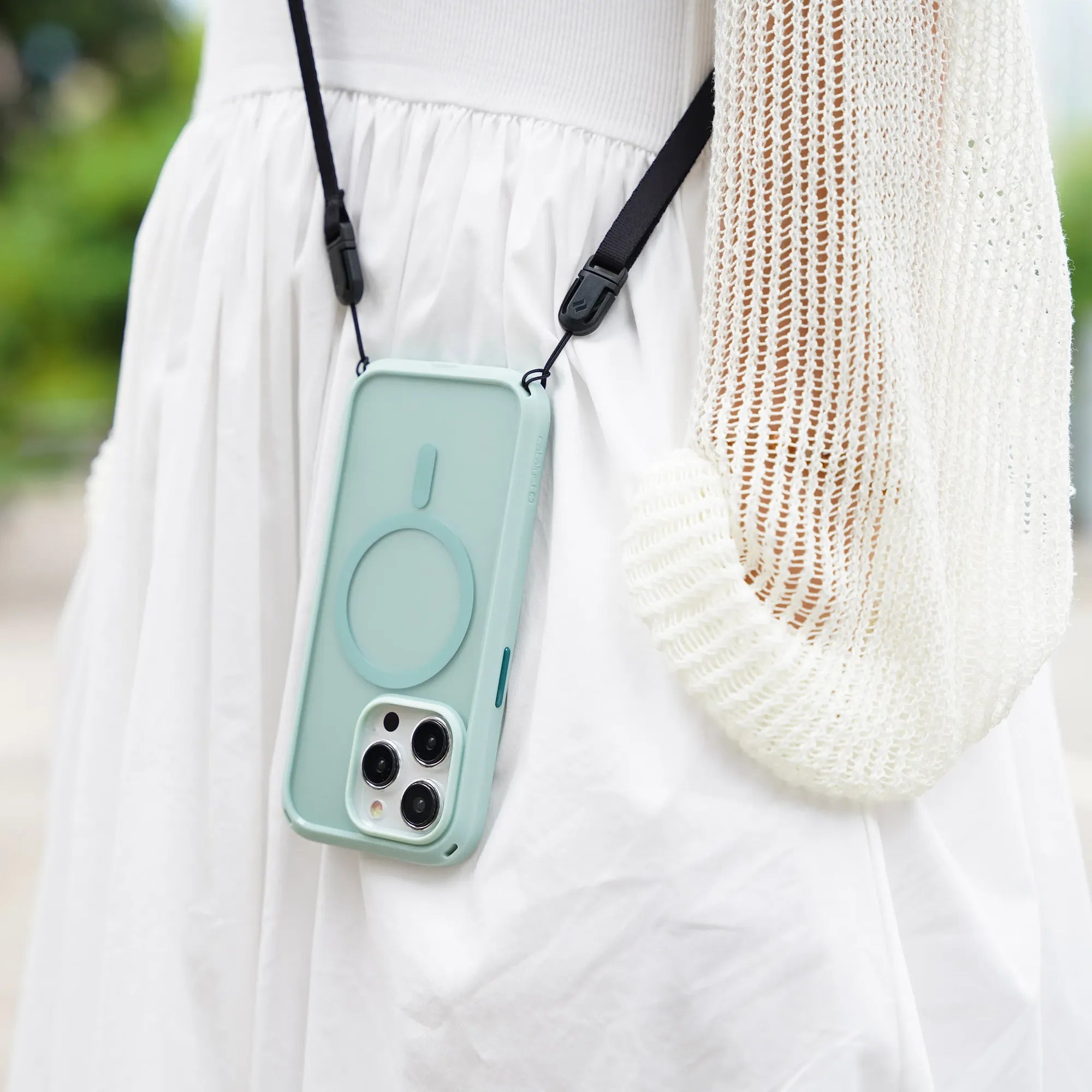 A person in a white skirt and cream knit sweater carries the Catalyst Case US iPhone 15 Pro - Influence Case (MagSafe Compatible) in light blue on a neck strap with black clips; the background shows blurred greenery.