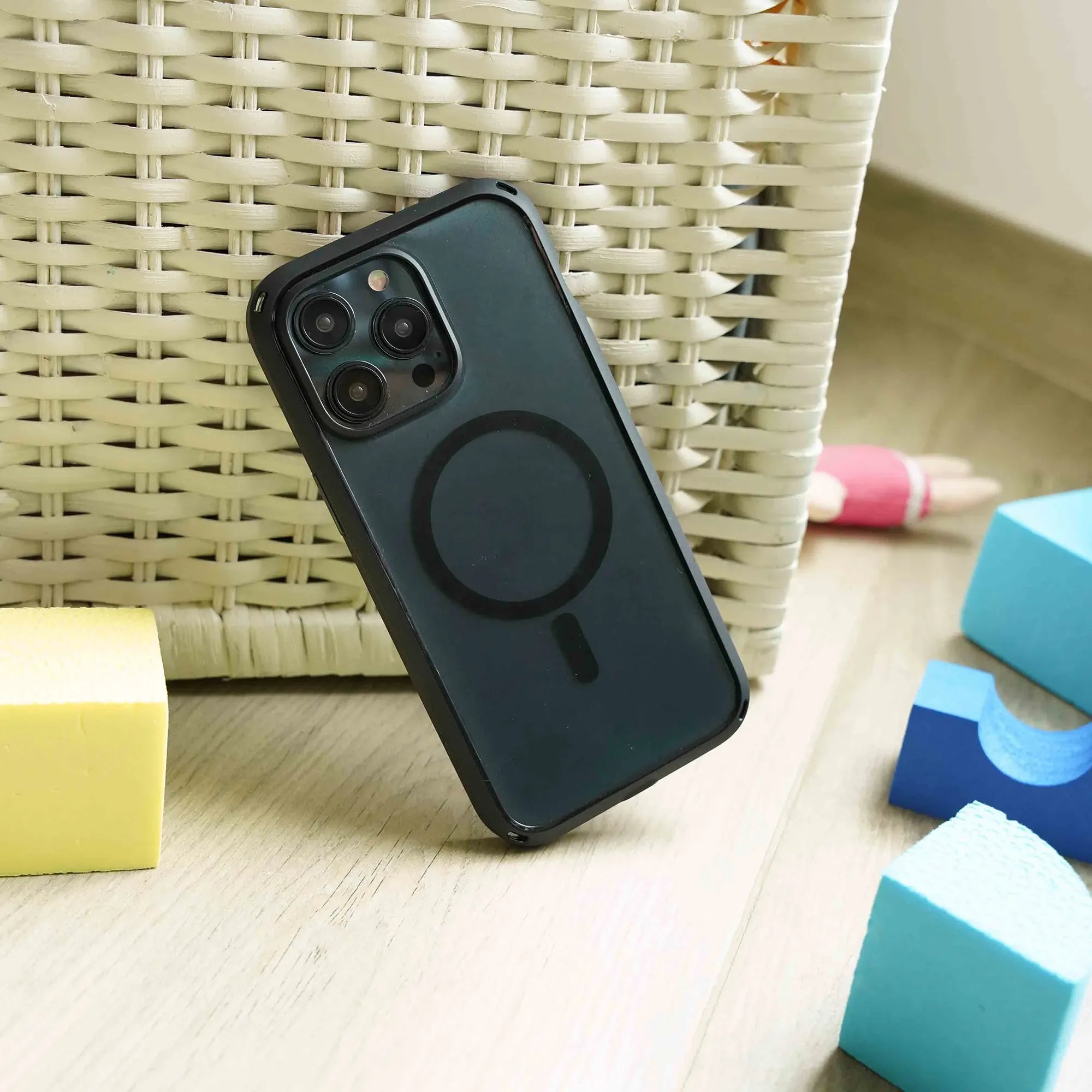 An iPhone 15 Pro in the black Catalyst Influence Case (MagSafe compatible) stands upright against a light wicker basket on a wooden floor, surrounded by colorful foam blocks in yellow, blue, and pink, softly lit by natural light.
