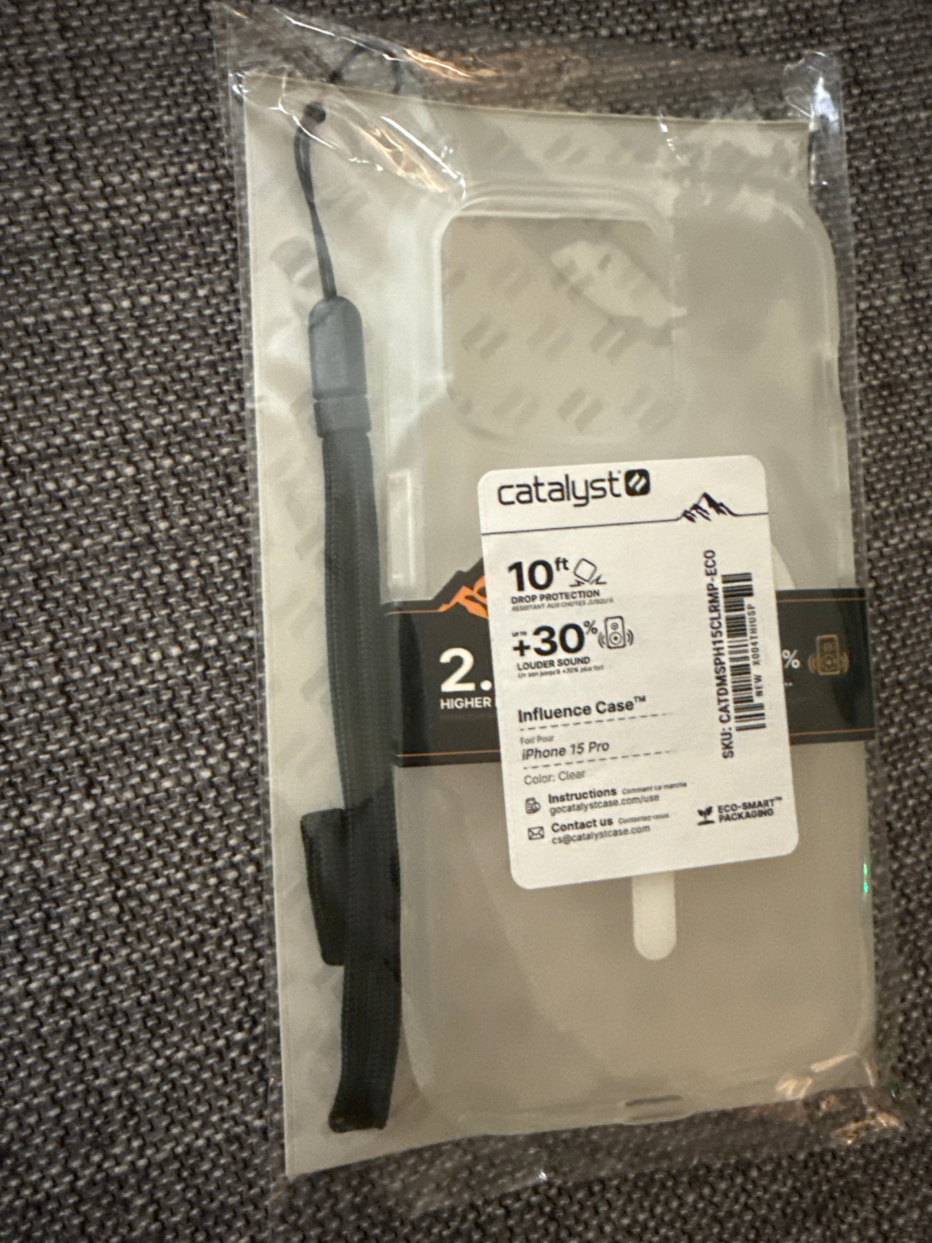 A packaged Catalyst Case US Influence Case, designed as a clear protective phone case for the iPhone 15 Pro with MagSafe compatibility, is displayed on a gray fabric surface. The packaging highlights military-grade drop protection up to 10 ft and includes a black wrist strap. A white label contains product information.
