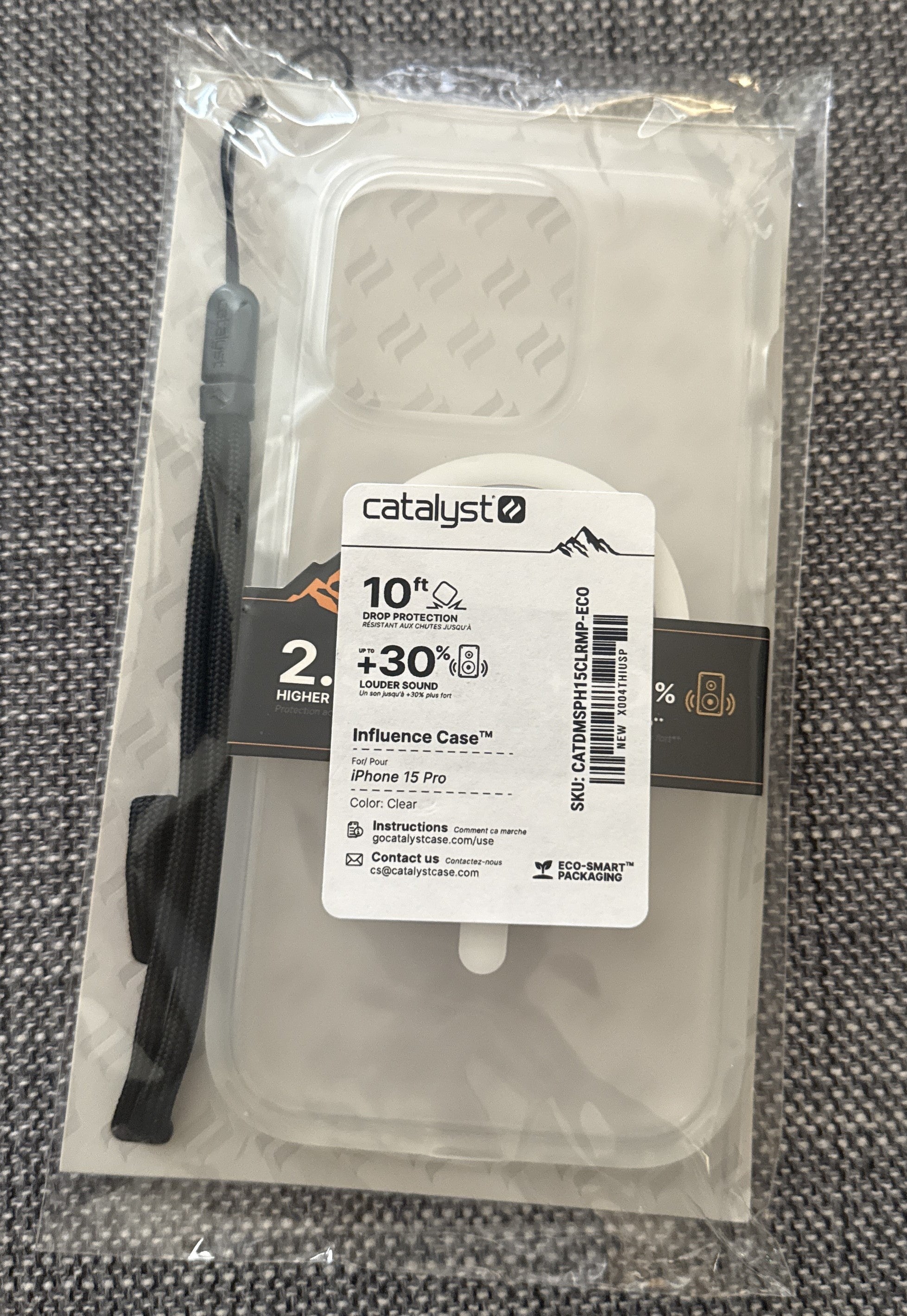 A Catalyst Case US iPhone 15 Pro - Influence Case (MagSafe Compatible) in Clear is sealed in transparent packaging. The package emphasizes military-grade drop protection up to 10 feet and claims 30% louder sound. A black wrist strap is attached on the left side of the packaging, which rests on a gray textured fabric surface.