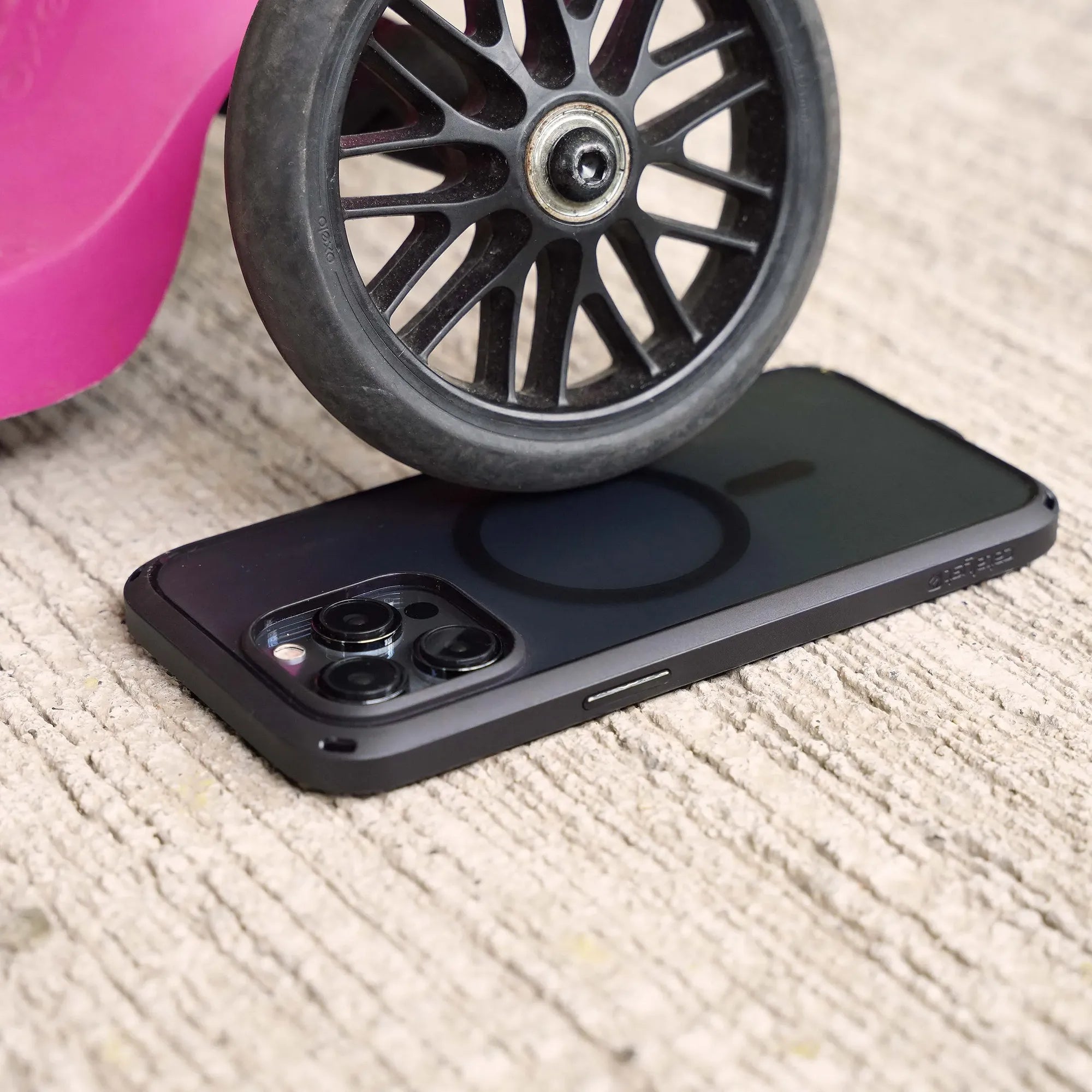 A black iPhone 14 Pro Max in a MagSafe-compatible Catalyst Influence Case lies face down on concrete, while a black plastic wheel from a pink toy presses on its back, highlighting the case’s durability with visible camera lenses and MagSafe ring.