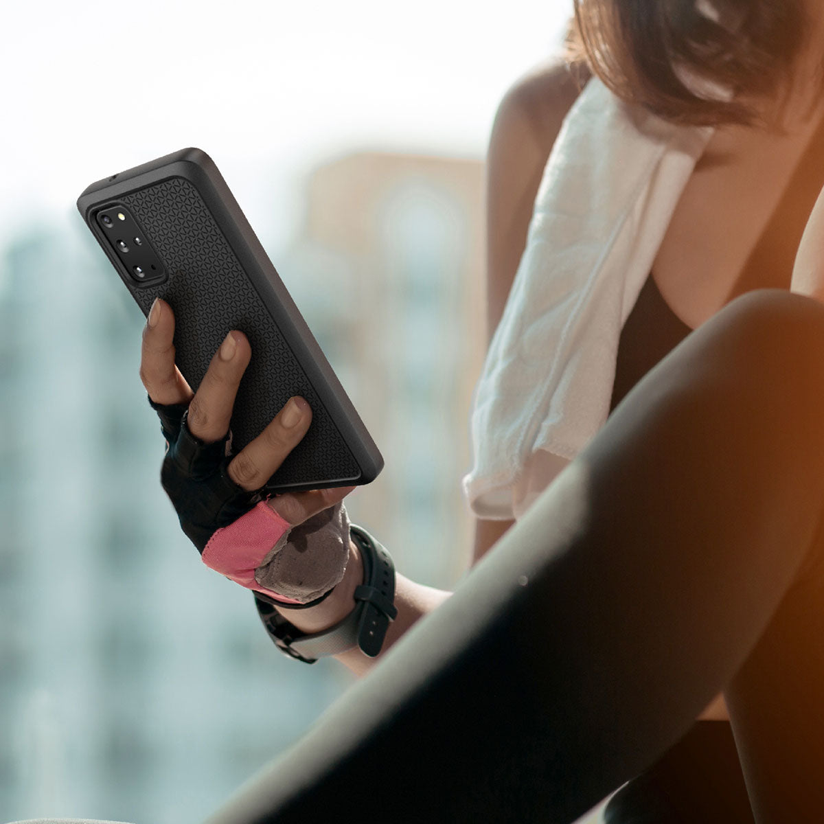 A woman in athletic wear sits outdoors, holding a Samsung Galaxy S20 Plus in a Catalyst Case US Impact Protection Case. She wears a smartwatch and fingerless glove, with a white towel over her shoulder; city buildings are blurred behind her.