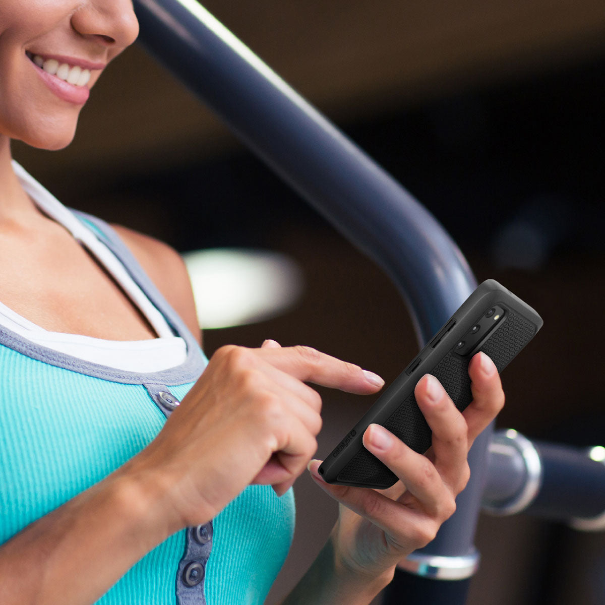 A smiling woman in a turquoise tank top uses her Samsung Galaxy S20 with a Catalyst Case US Impact Protection Case near gym equipment, her finger poised above the screen. The softly blurred background suggests an indoor workout setting.