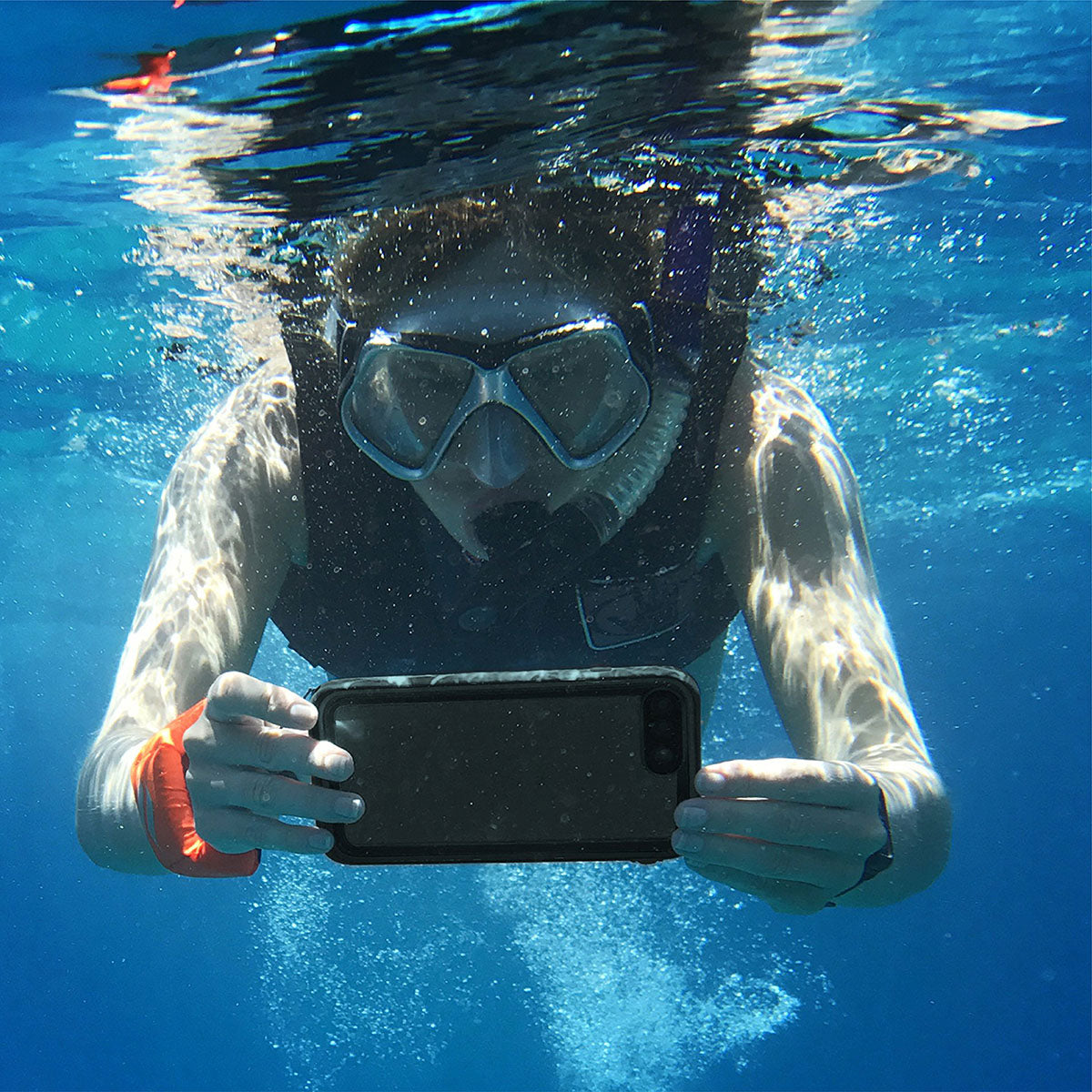 Underwater, a person in snorkeling gear holds an iPhone 8 Plus/7 Plus secured in a Catalyst Case US Waterproof Case. Sunlight shimmers on their arms as bubbles rise behind them in the clear blue water.